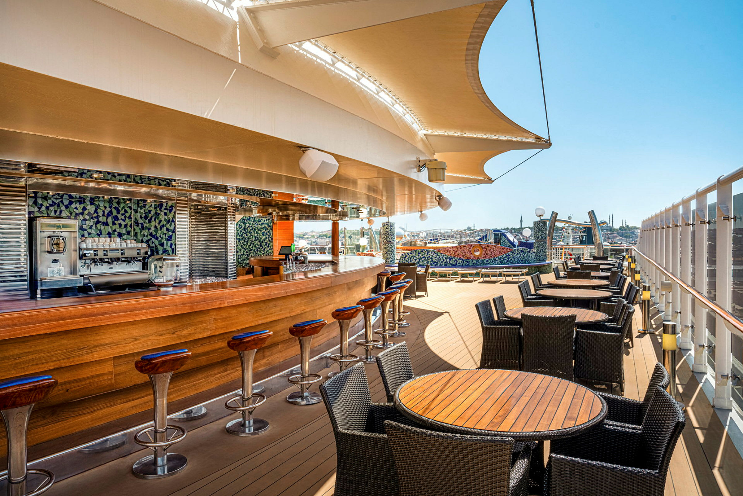 Bilde av en utendørsbar med treinnredning og sitteplasser omgitt av bord og stoler, under en stor seilduk med utsikt over byens skyline. (AI-generert tekst)