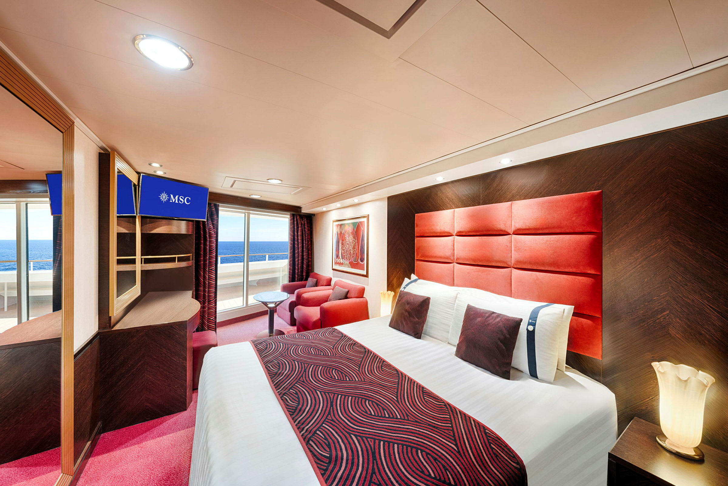 Et moderne hotellrom med en dobbeltseng, røde mønstrede tekstiler, røde lenestoler, et lite bord og en balkong med utsikt over havet. (AI-generert tekst)
