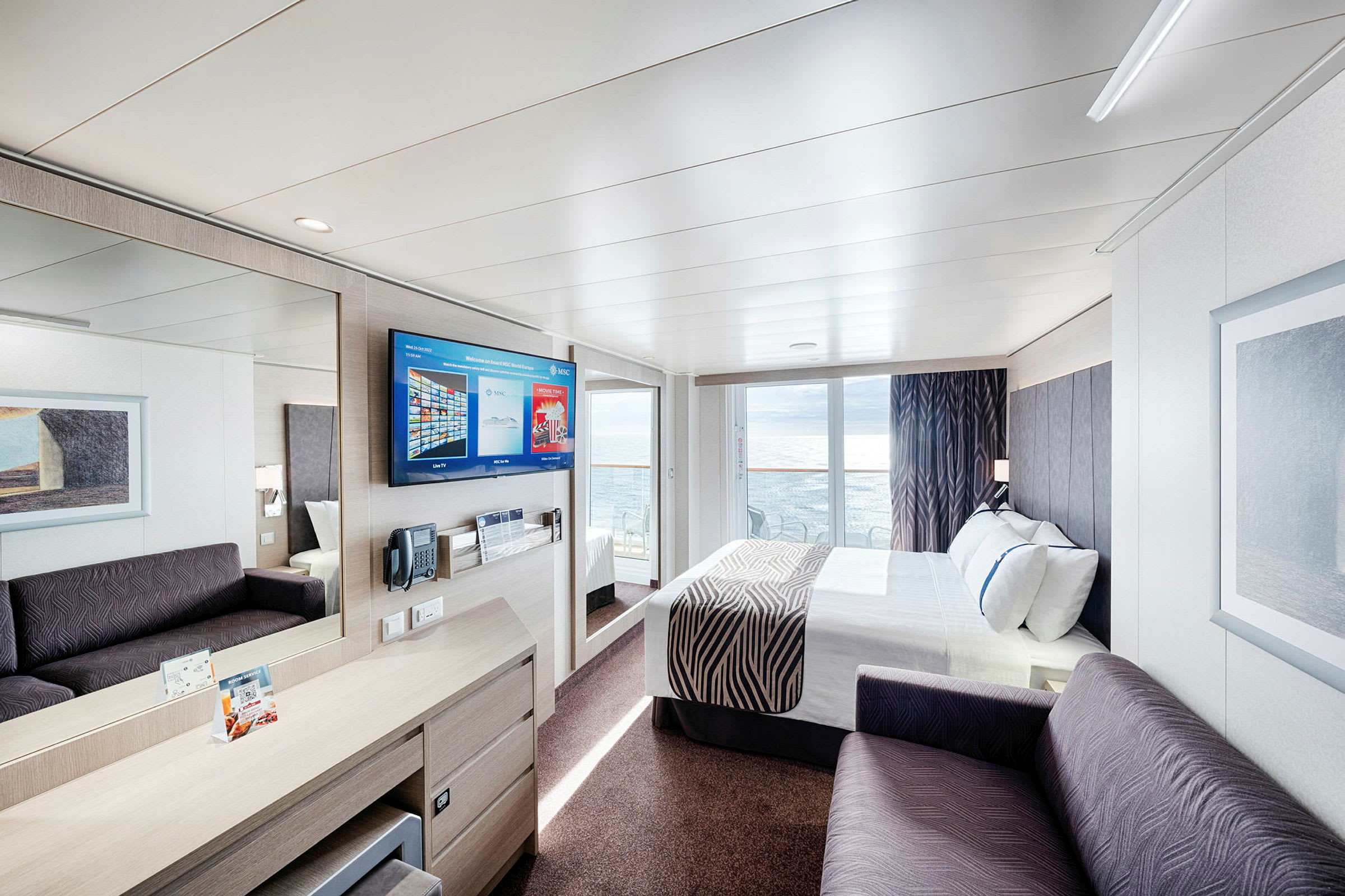 Et moderne hotellrom med seng, sofa og en TV på veggen. Glassdører fører ut til en balkong med havutsikt. (AI-generert tekst)