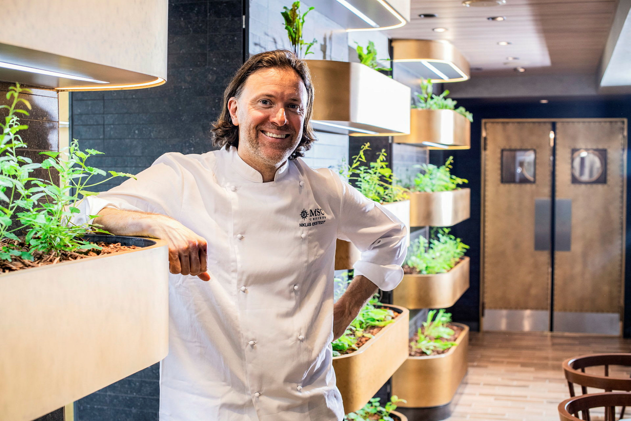 Kokken står ved siden av planteinstallasjoner i en restaurantmiljø. (AI-generert tekst)