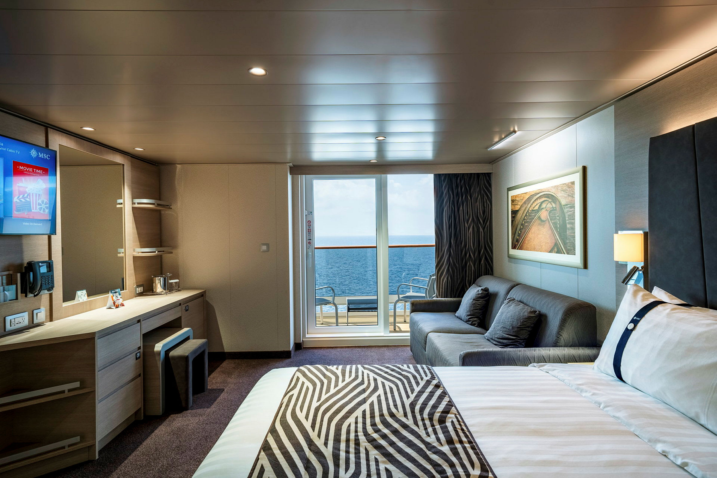 En interiør av et hotellrom med seng, sofa, skrivebord og en balkong som har utsikt over havet. (AI-generert tekst)