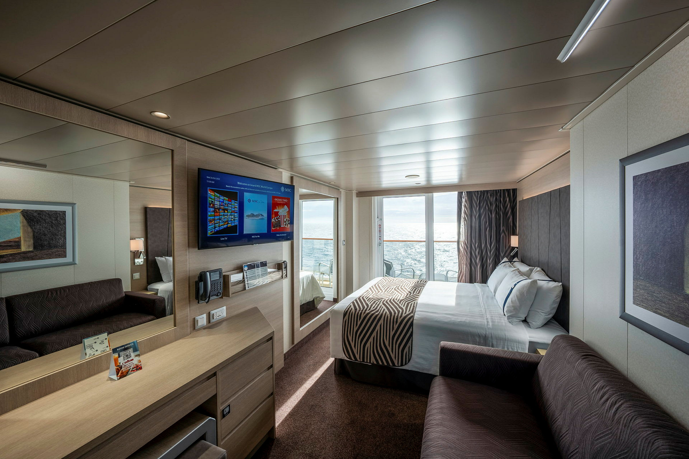 Moderne innredet hotellrom med dobbeltseng, sofa, skrivebord og veggmontert TV. Stor glassdør leder til balkong med havutsikt. (AI-generert tekst)
