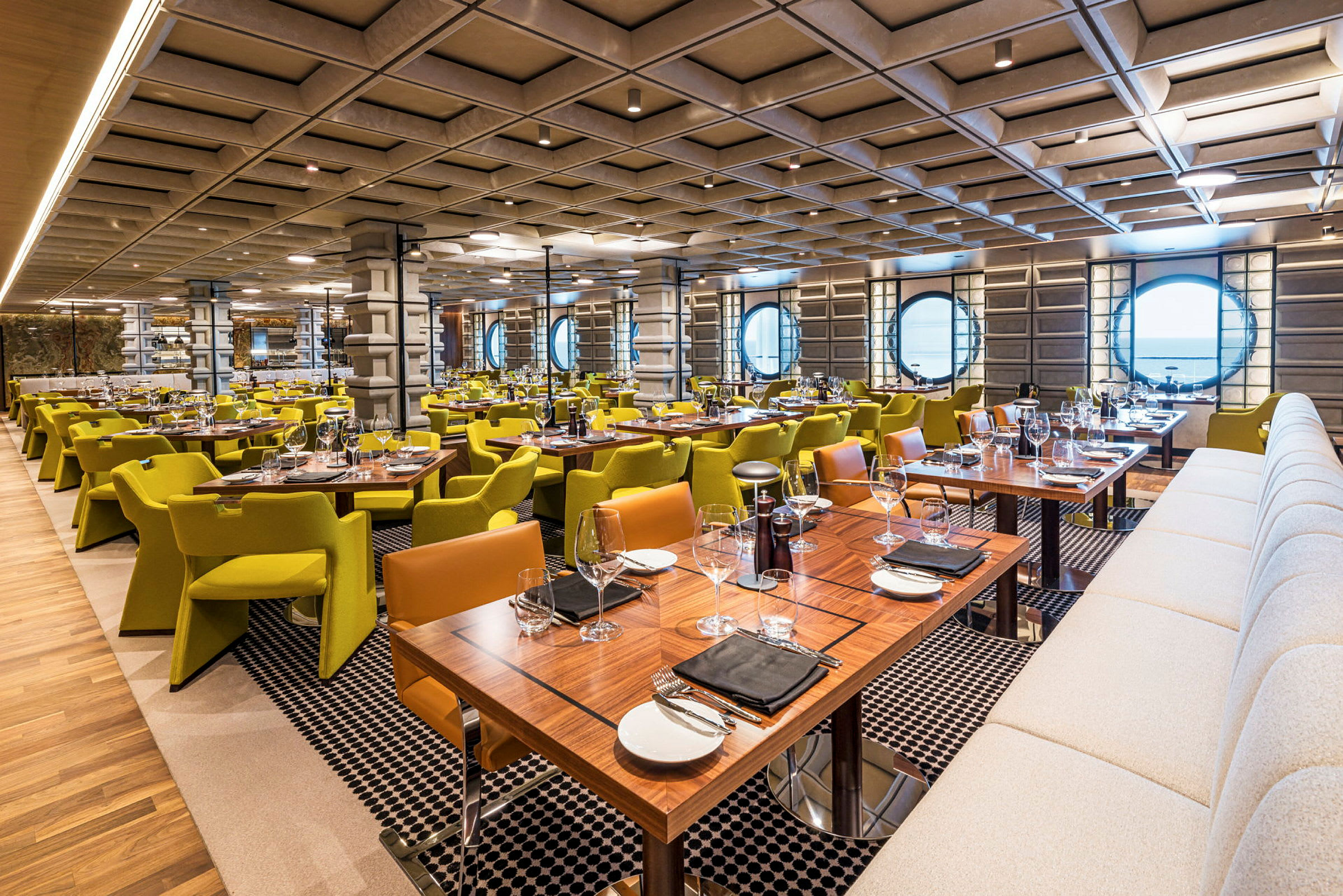 Interiøret til en moderne restaurant med mange dekkede bord, grønne stoler og en buet vegg med runde vinduer. (AI-generert tekst)