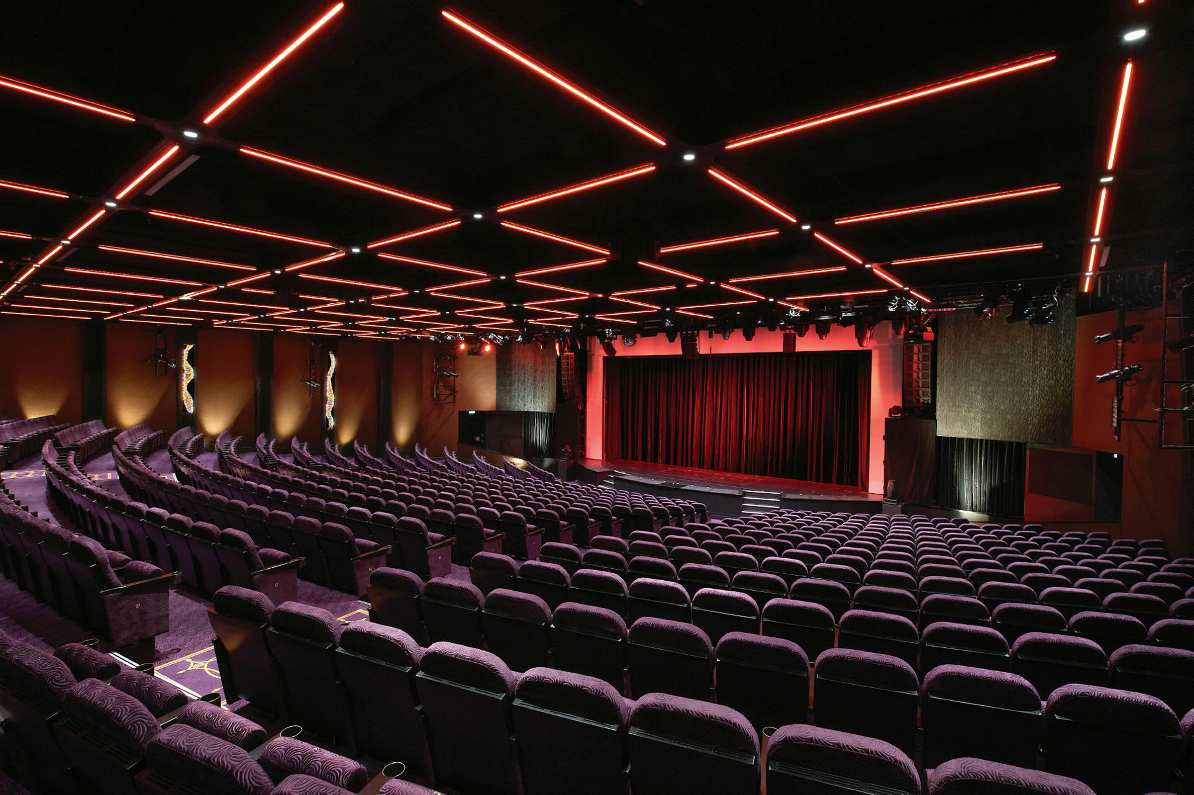 En teatersal med rader av lilla stoler og et belyst tak med røde linjer. Scenen er kledd med røde gardiner. (AI-generert tekst)