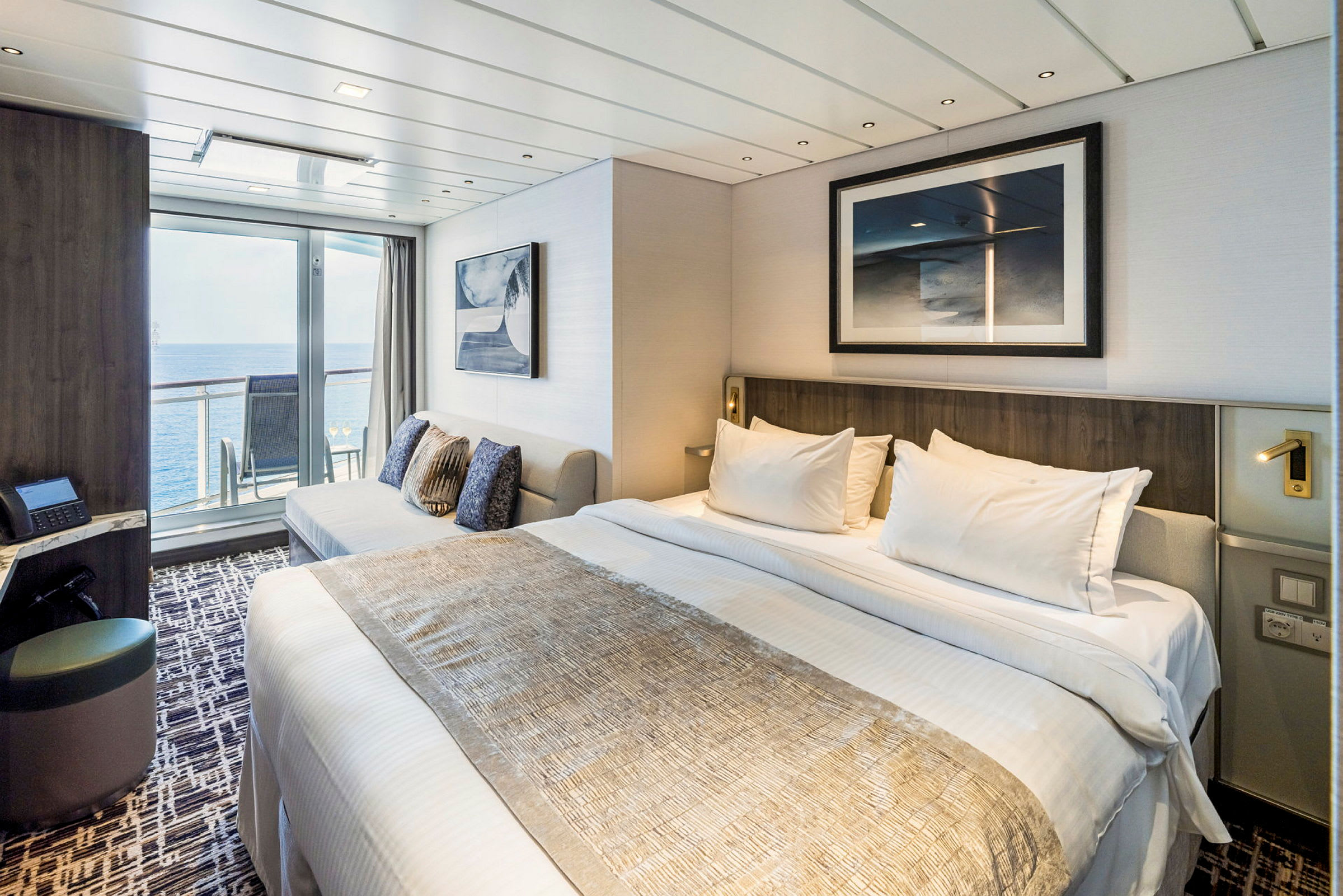 Et moderne hotellrom med en oppredd seng, en sofa og glassdører som fører ut til en balkong med utsikt over havet. (AI-generert tekst)