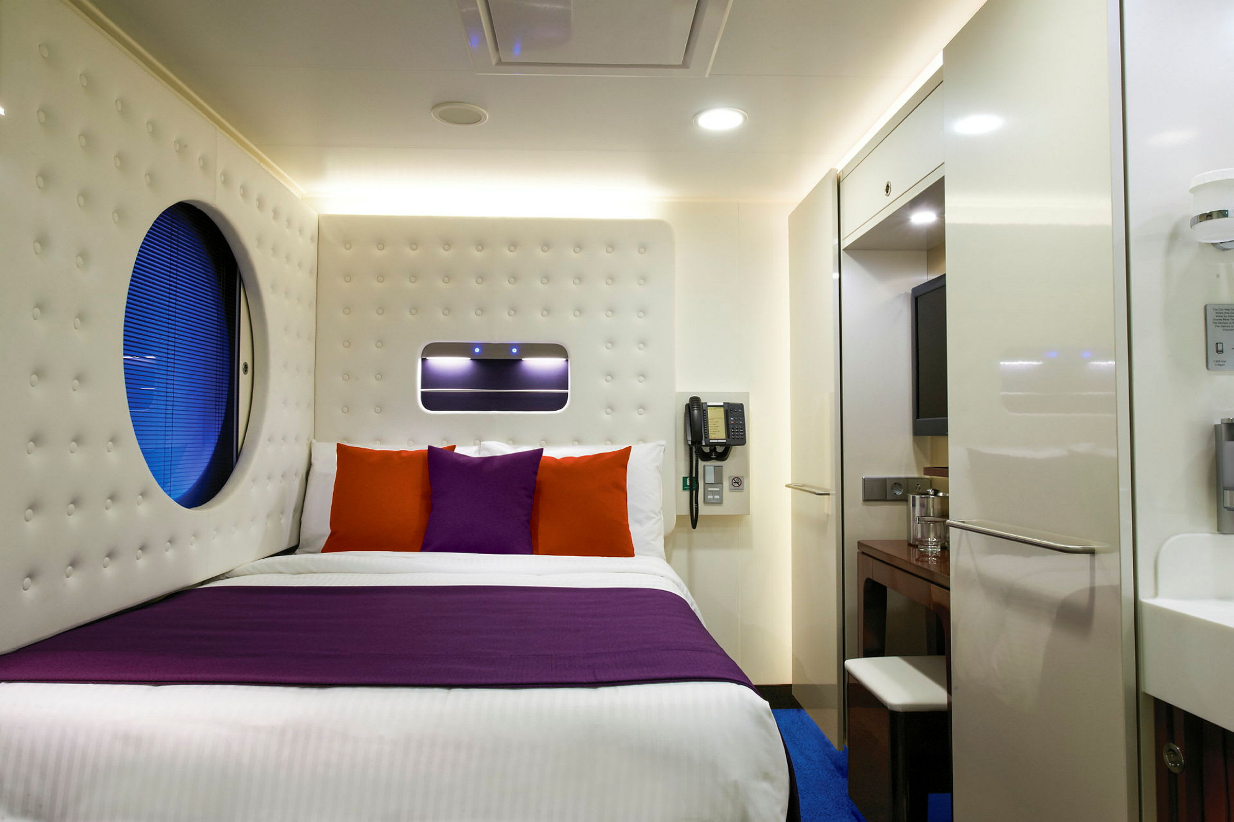 Et moderne hotellrom med en seng som har lilla og oransje puter. (AI-generert tekst)