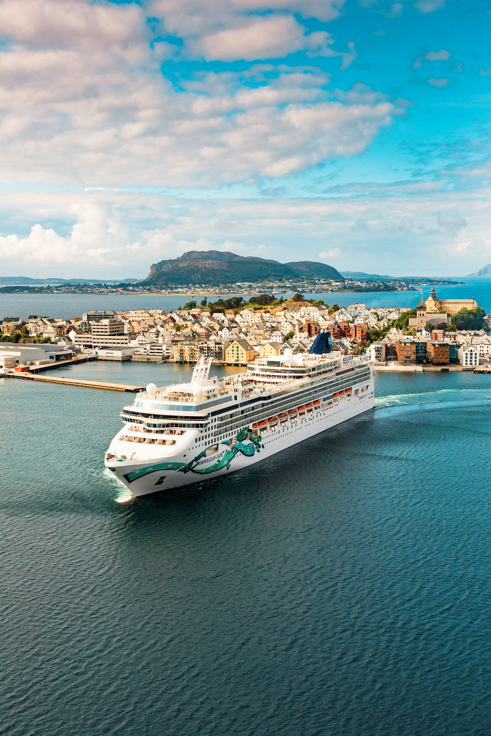 Et stort cruiseskip passerer nær en by ved vannet med fjell i bakgrunnen. (AI-generert tekst)