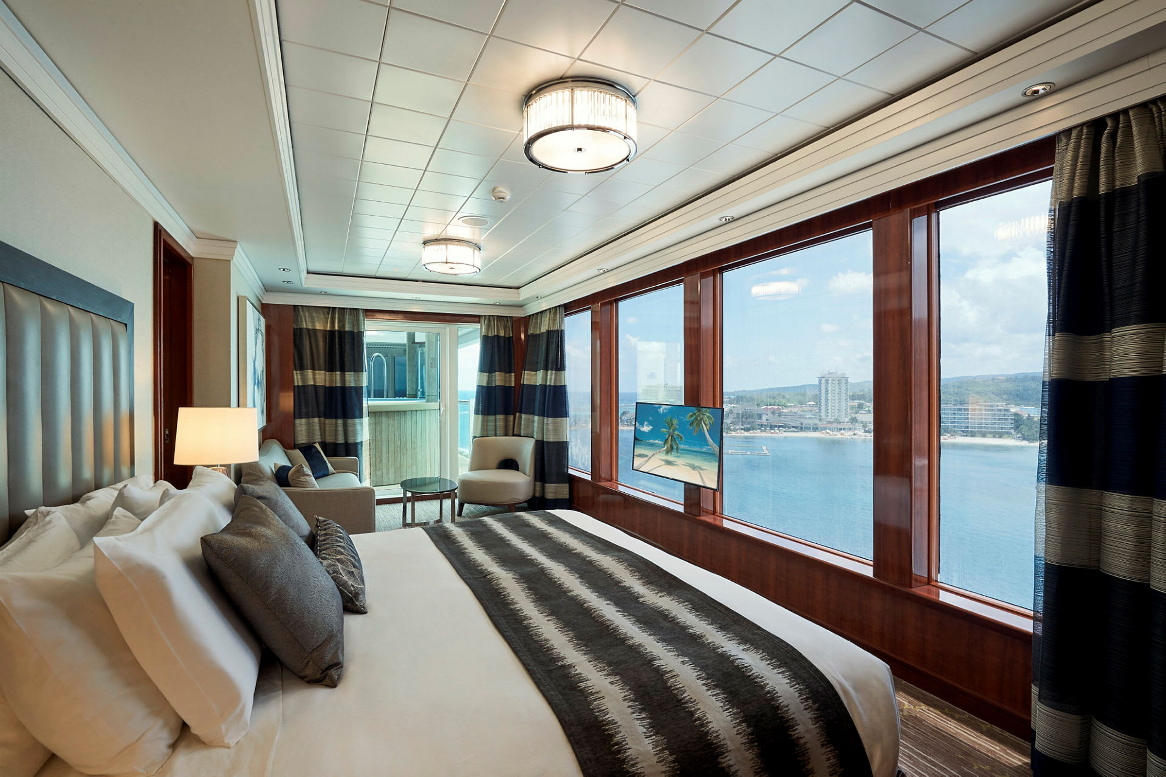 Et elegant hotellrom med en stor seng, to lenestoler og store vinduer som viser utsikt over vannet og en kystlinje. (AI-generert tekst)