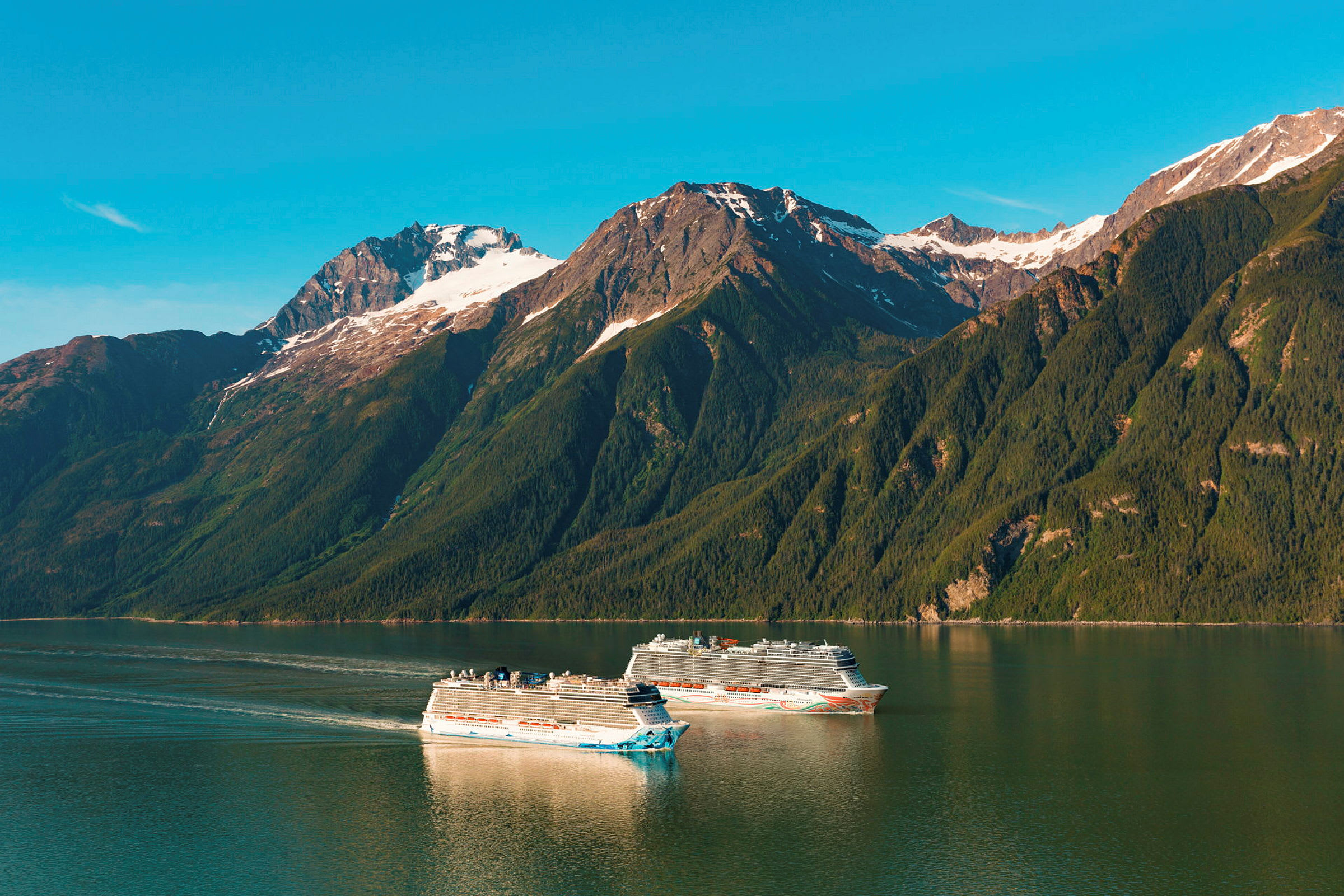 To cruiseskip seiler i en rolig fjord med skogkledde fjell og snødekte topper i bakgrunnen. (AI-generert tekst)