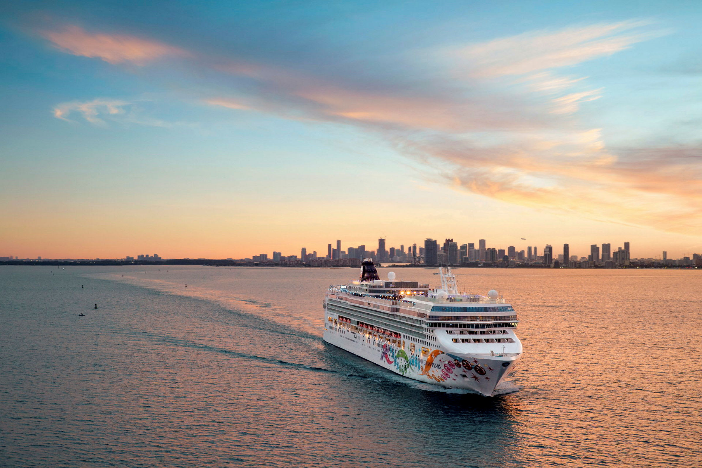 Et cruiseskip seiler på havet med en bysilhuett i bakgrunnen ved solnedgang. (AI-generert tekst)