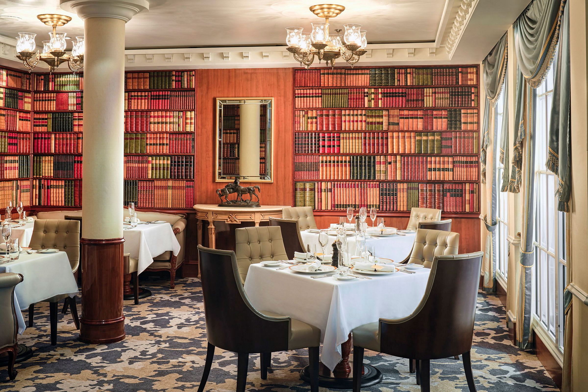 En elegant restaurant med bord dekket med hvite duker, omgitt av bokhyller samt stort vindu og rike gardiner. (AI-generert tekst)