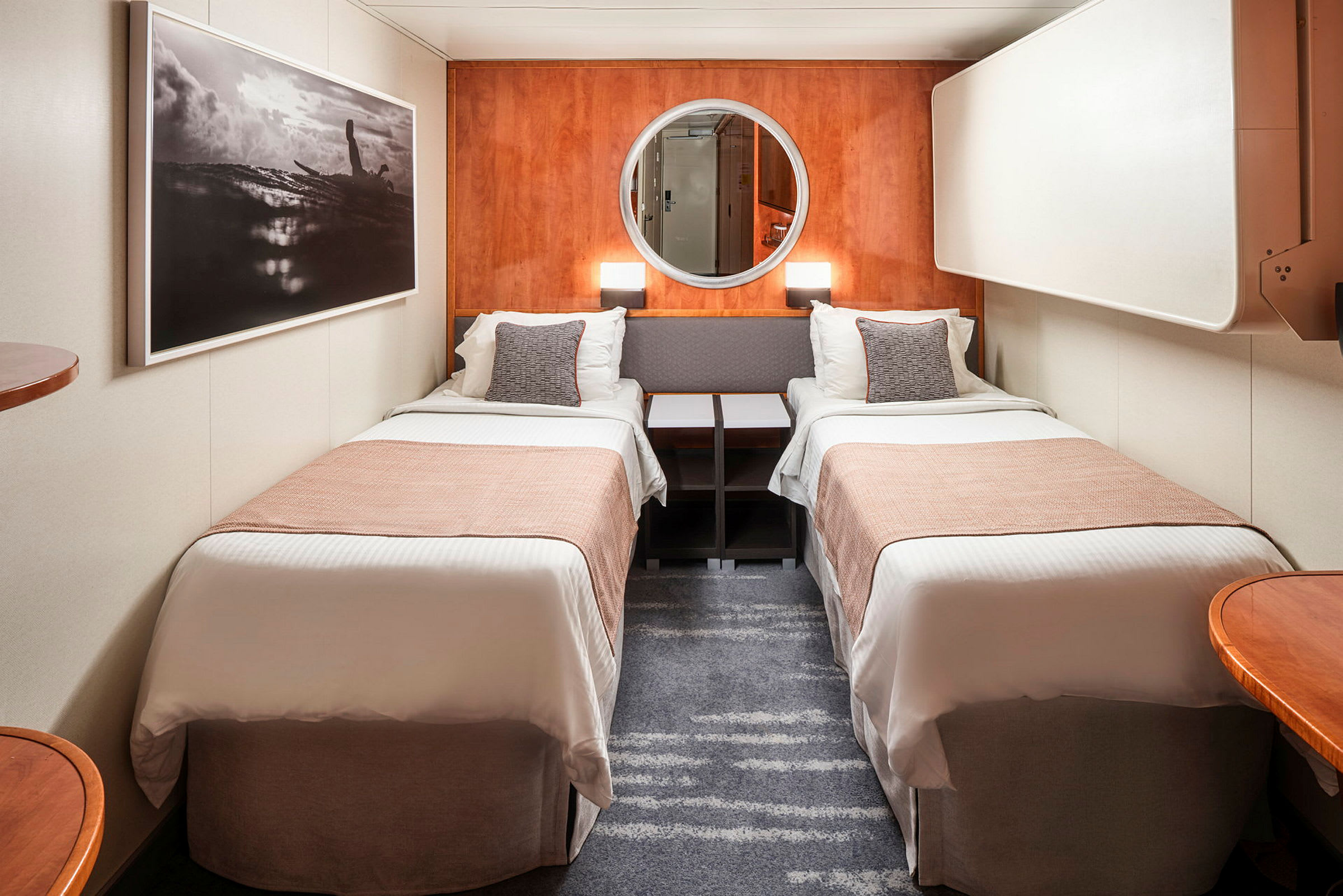 Et hotellrom med to enkeltsenger adskilt av et nattbord. Veggene er dekorert med et stort maleri og et rundt speil. (AI-generert tekst)