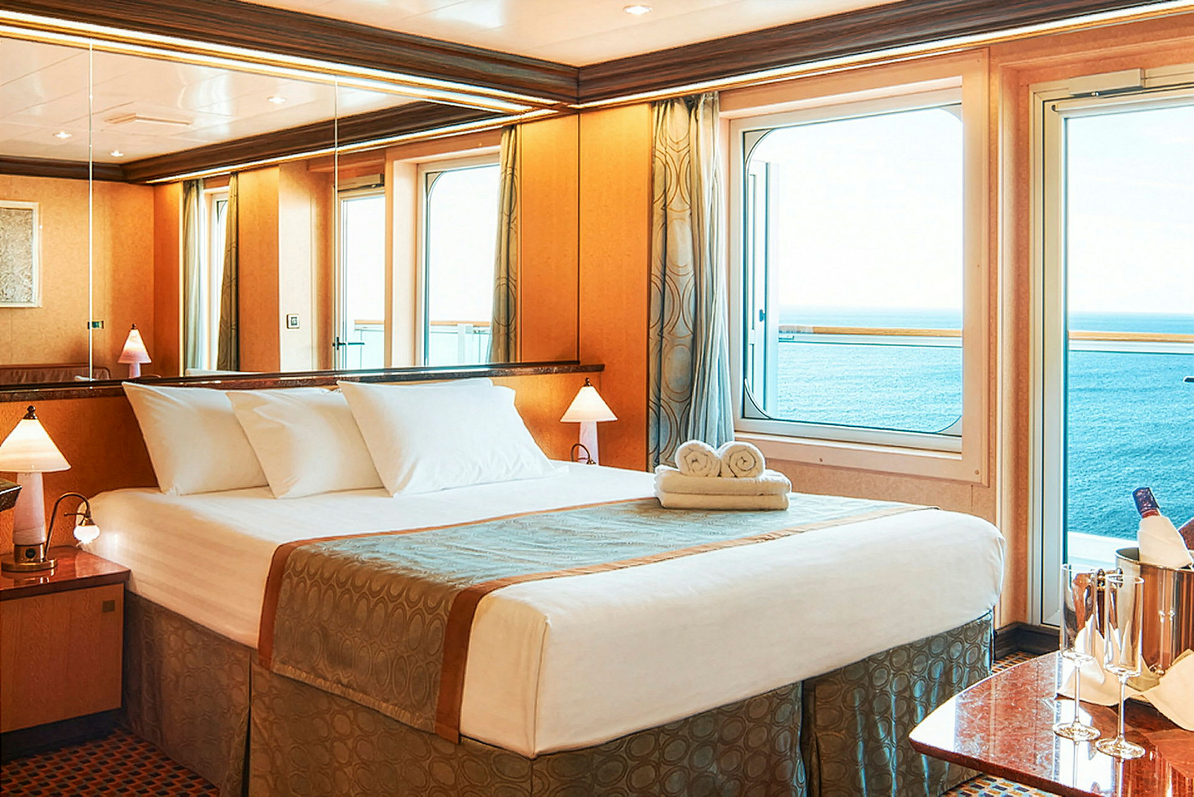 Et hotellrom med en oppredd seng, utsikt over havet gjennom vinduer og bord med champagneglass. (AI-generert tekst)