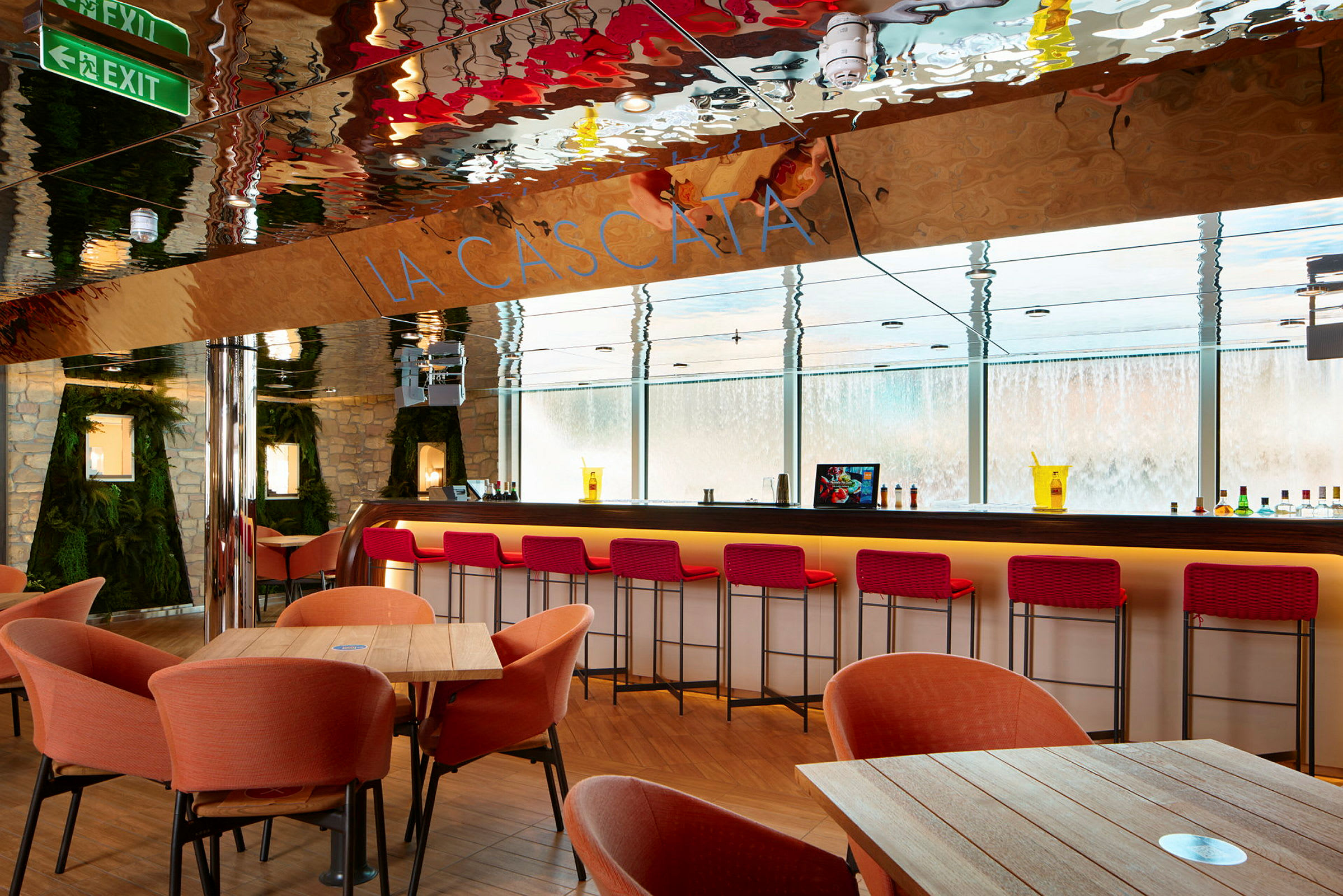 En moderne bar med røde barkrakker, oransje lenestoler og trebord. Veggene er dekorert med stein og grønt. Glassvinduer viser en vanninstallasjon utenfor. (AI-generert tekst)