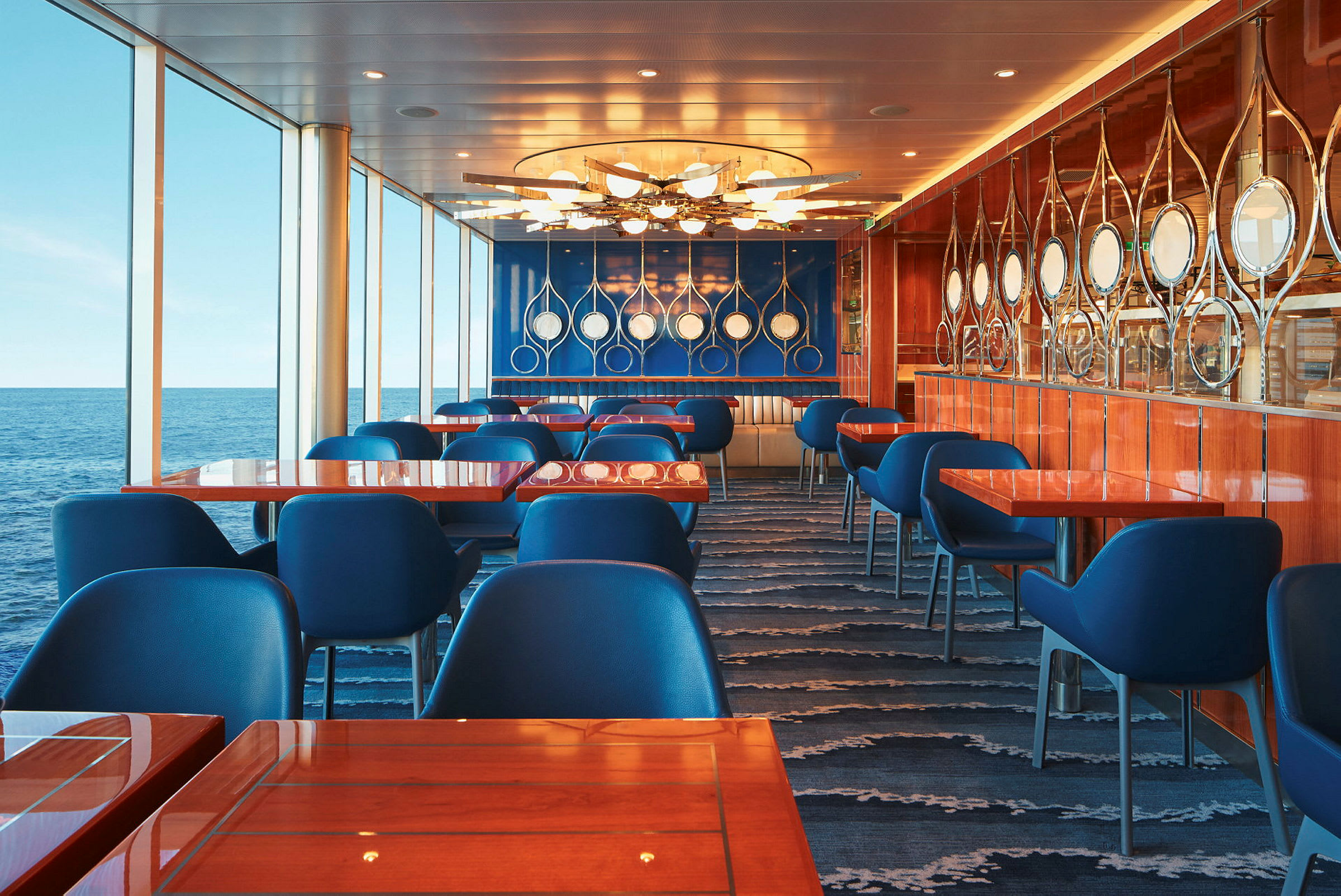 Restaurant med blå stoler, bord med treoverflate og panoramavinduer mot havet. (AI-generert tekst)