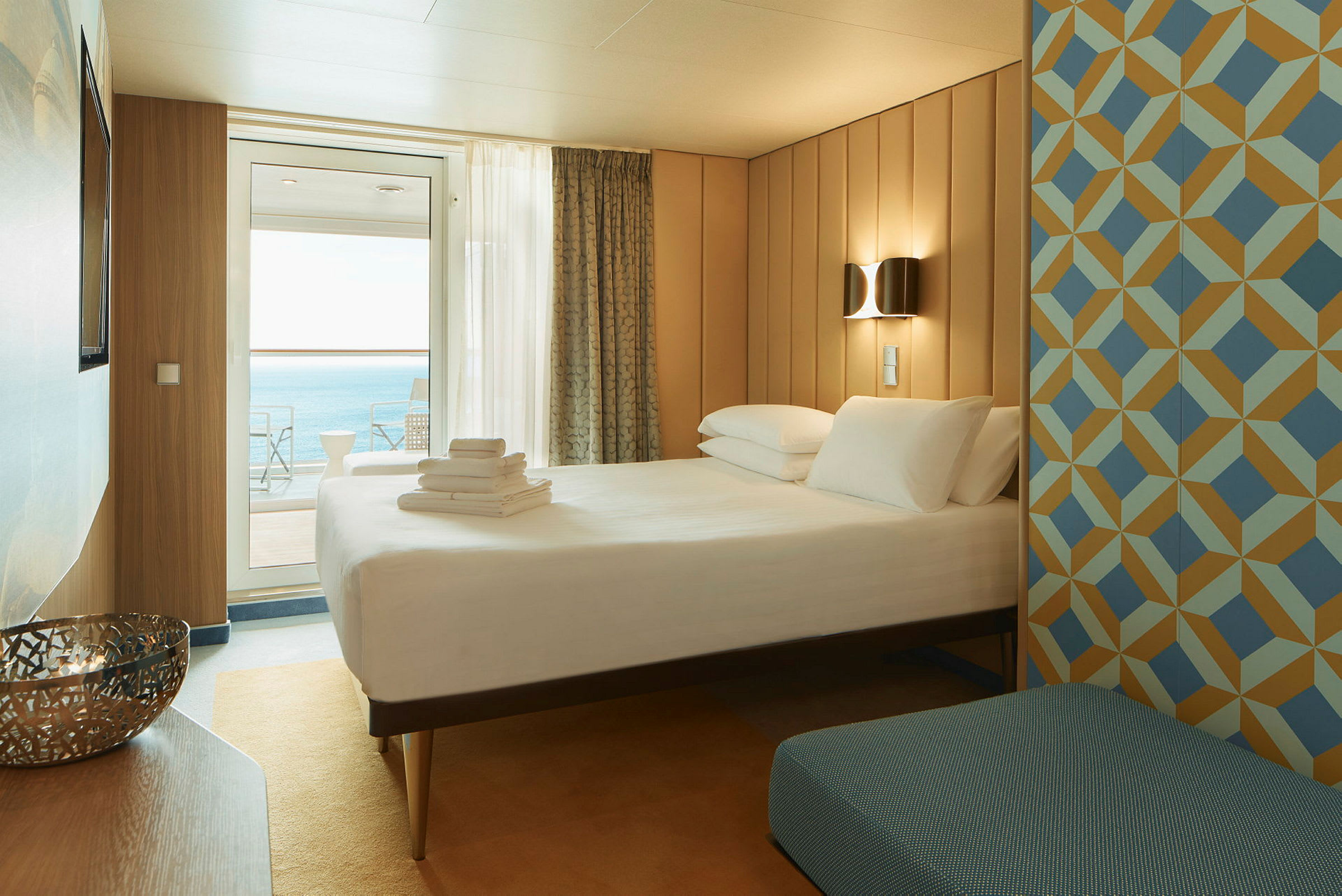 Et hotellrom med en dobbeltseng, geometriskmønstrede veggpaneler og et vindu med utsikt mot havet. (AI-generert tekst)