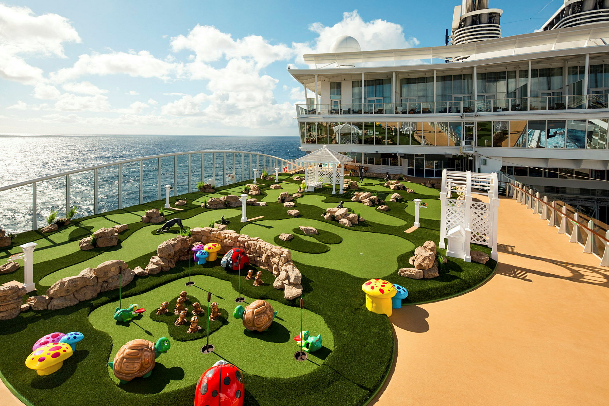 Bildet viser en minigolfbane ombord på et cruiseskip. Banen har kunstgress med steinutsmykninger og fargerike dekorasjoner, samt havet i bakgrunnen. (AI-generert tekst)