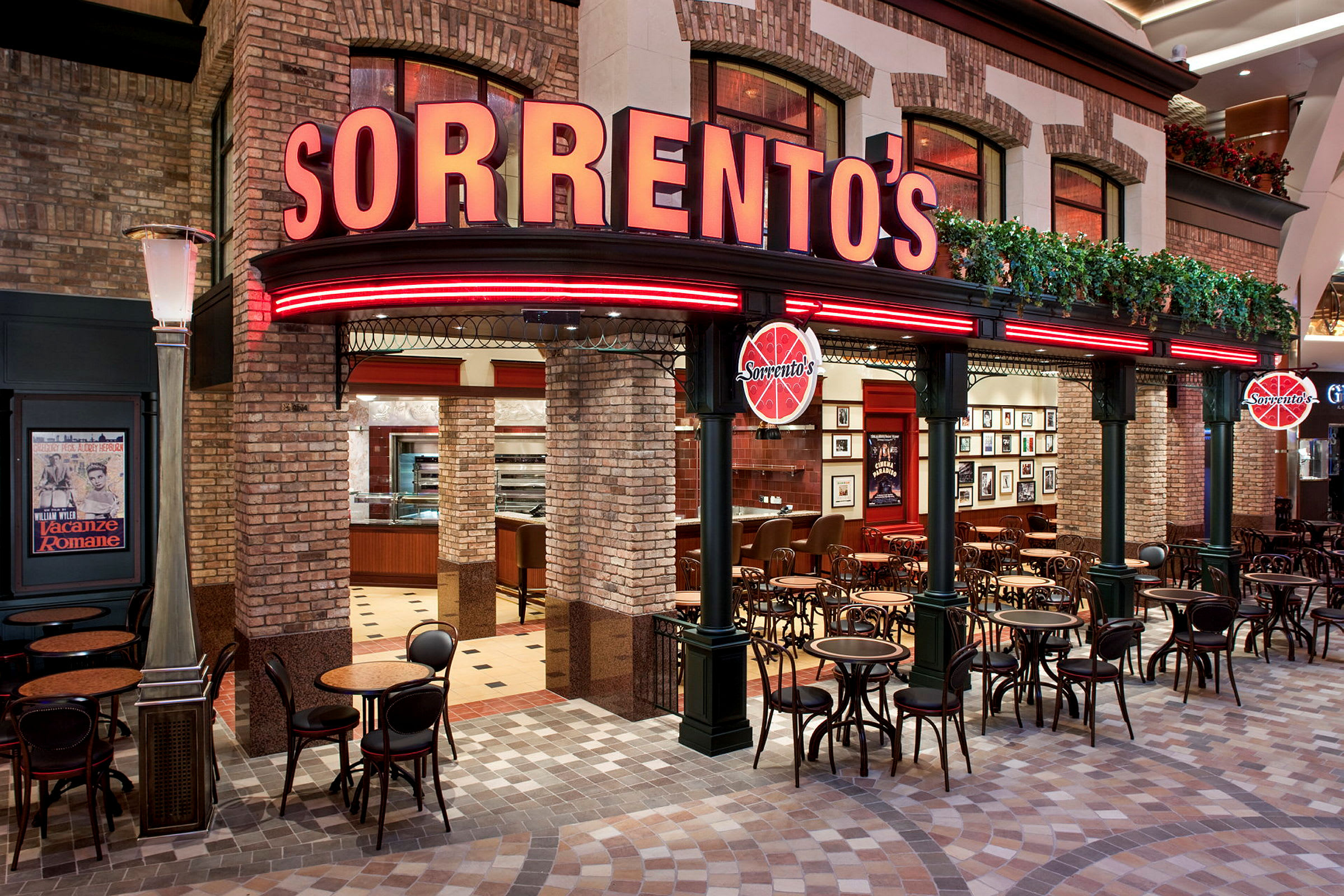 Eksteriør av en restaurant med navnet "Sorrento's". Bygningen har teglvegger og røde neonskilt. Foran restauranten er det små bord og stoler. (AI-generert tekst)