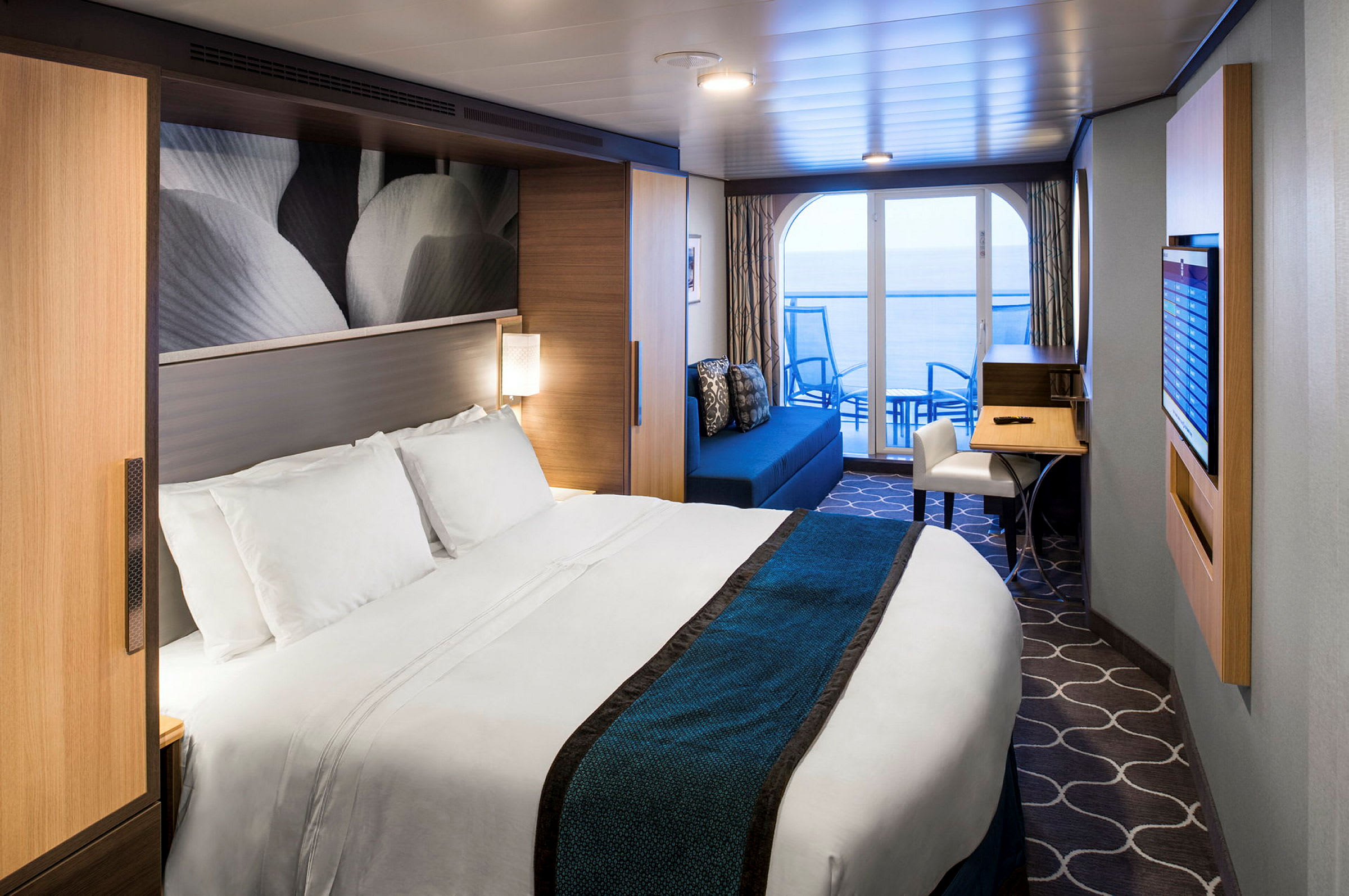 Et moderne hotellrom med en stor seng, dekorert med hvite og blå sengeklær. Rommet har en sittegruppe og skrivebord, samt en balkong som vender mot havet. (AI-generert tekst)