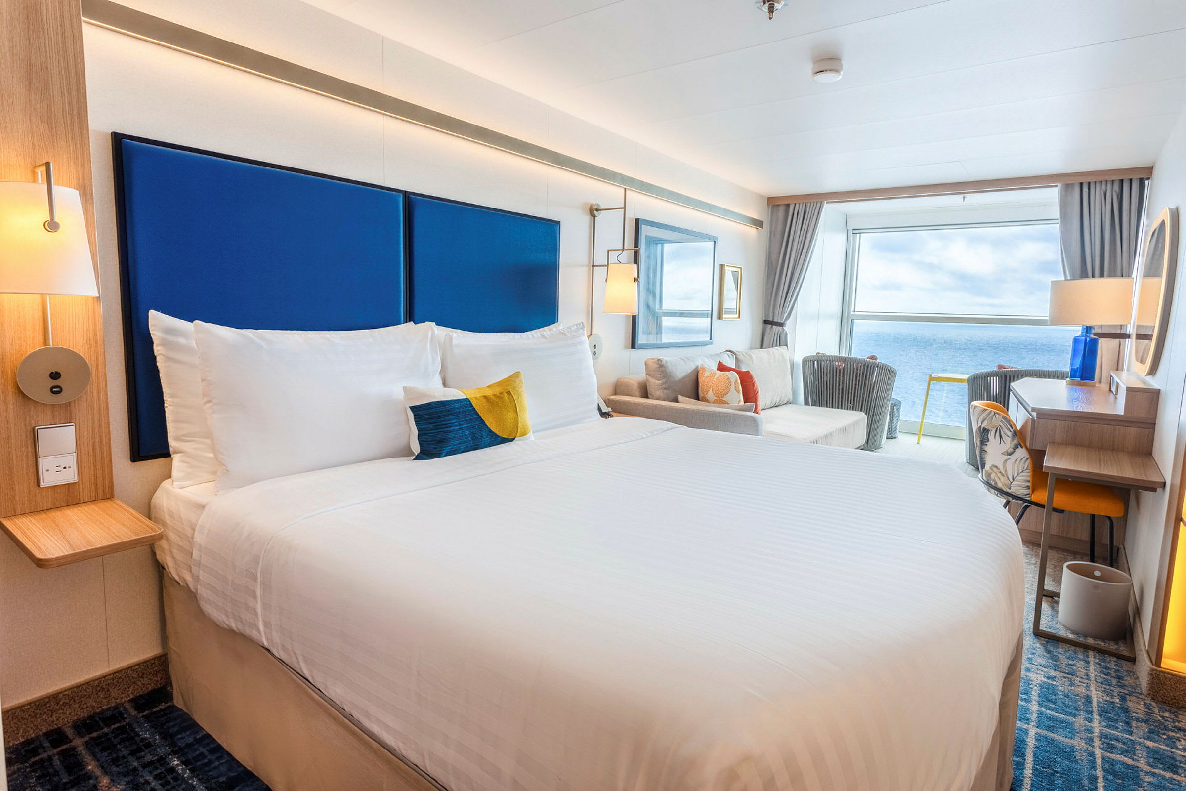 Et lyst hotellrom med en stor seng, sittegruppe og utsikt over havet. (AI-generert tekst)