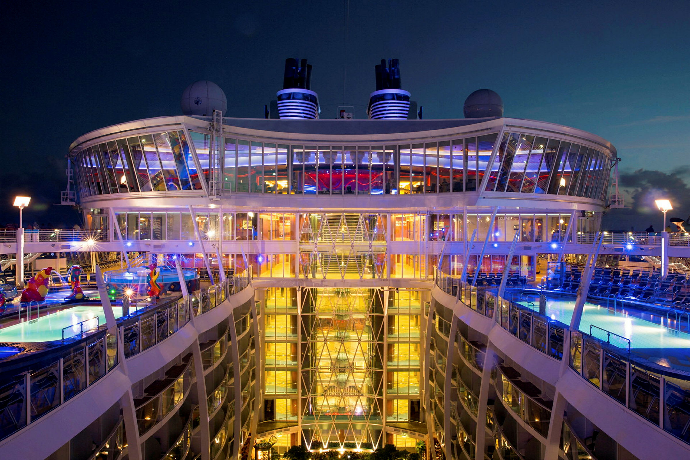 En kveldsscene som viser et opplyst cruiseskip med basseng og glassstruktur. (AI-generert tekst)