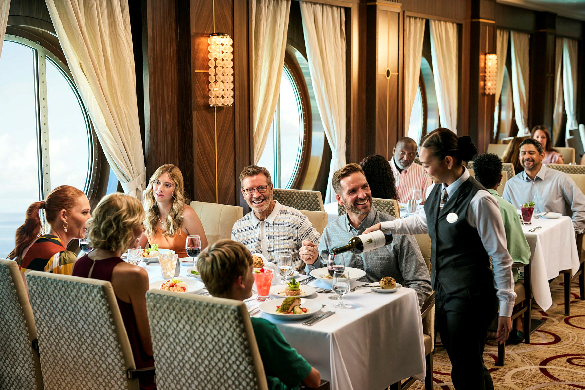 En elegant restaurant med et vindu med utsikt over havet, hvor mennesker nyter et måltid og en kelner serverer vin. (AI-generert tekst)