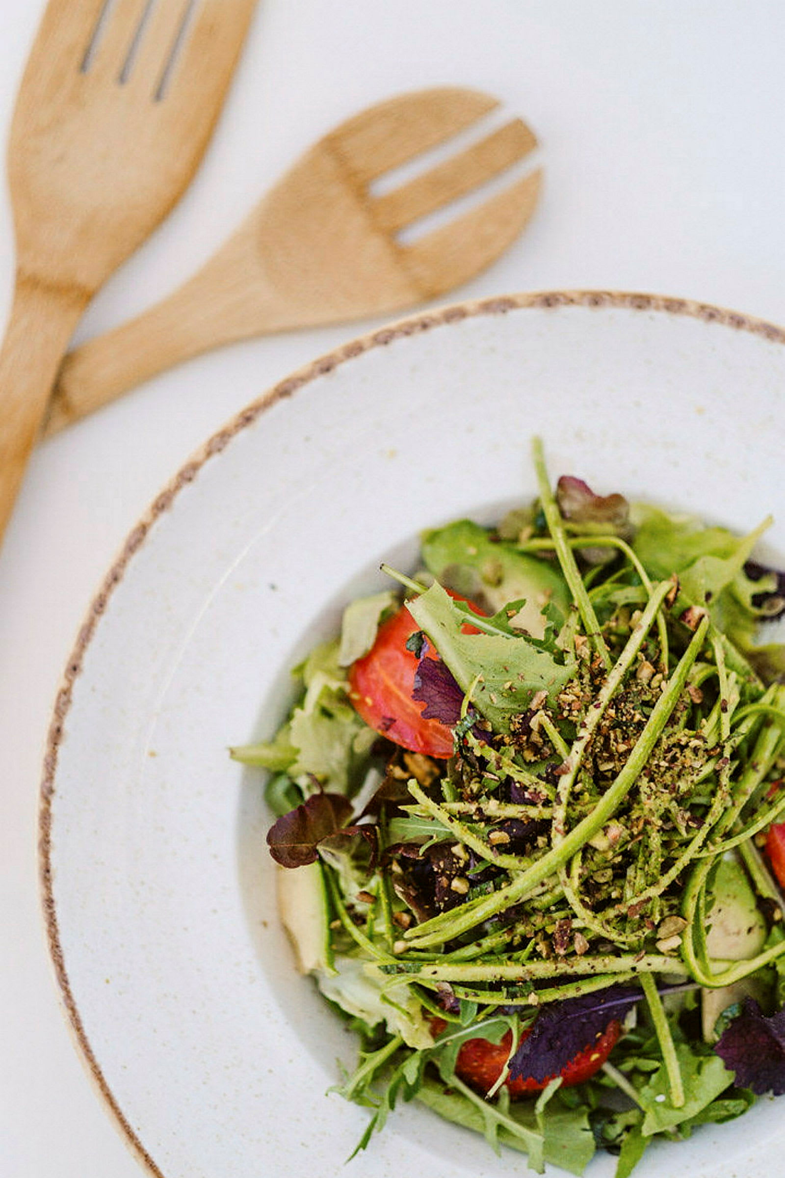 En tallerken med en fargerik salat som inneholder bladgrønn, tomater og avokado, med grovhakkede nøtter på toppen. To trebestikk ligger ved siden av. (AI-generert tekst)