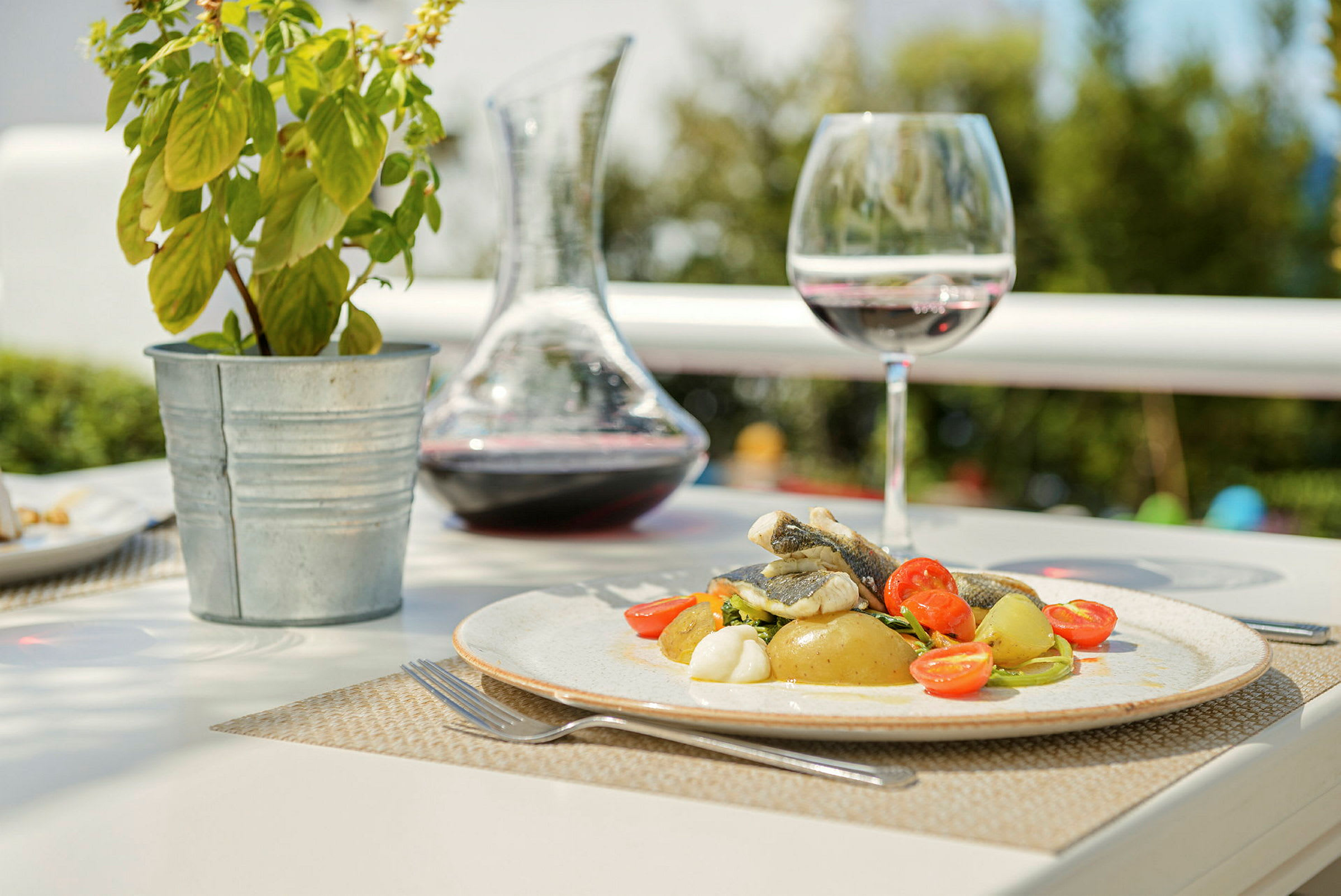 En tallerken med mat, bestående av fisk, poteter og cherrytomater, står på et bord sammen med et vinglass, en karaffel med vin og en potteplante. (AI-generert tekst)