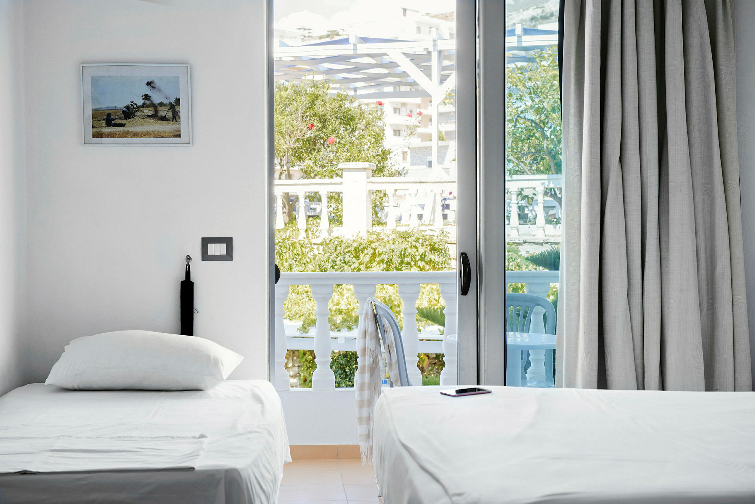 Et lyst hotellrom med to enkeltsenger, en balkong med utsikt til grøntområder og utemøbler. (AI-generert tekst)