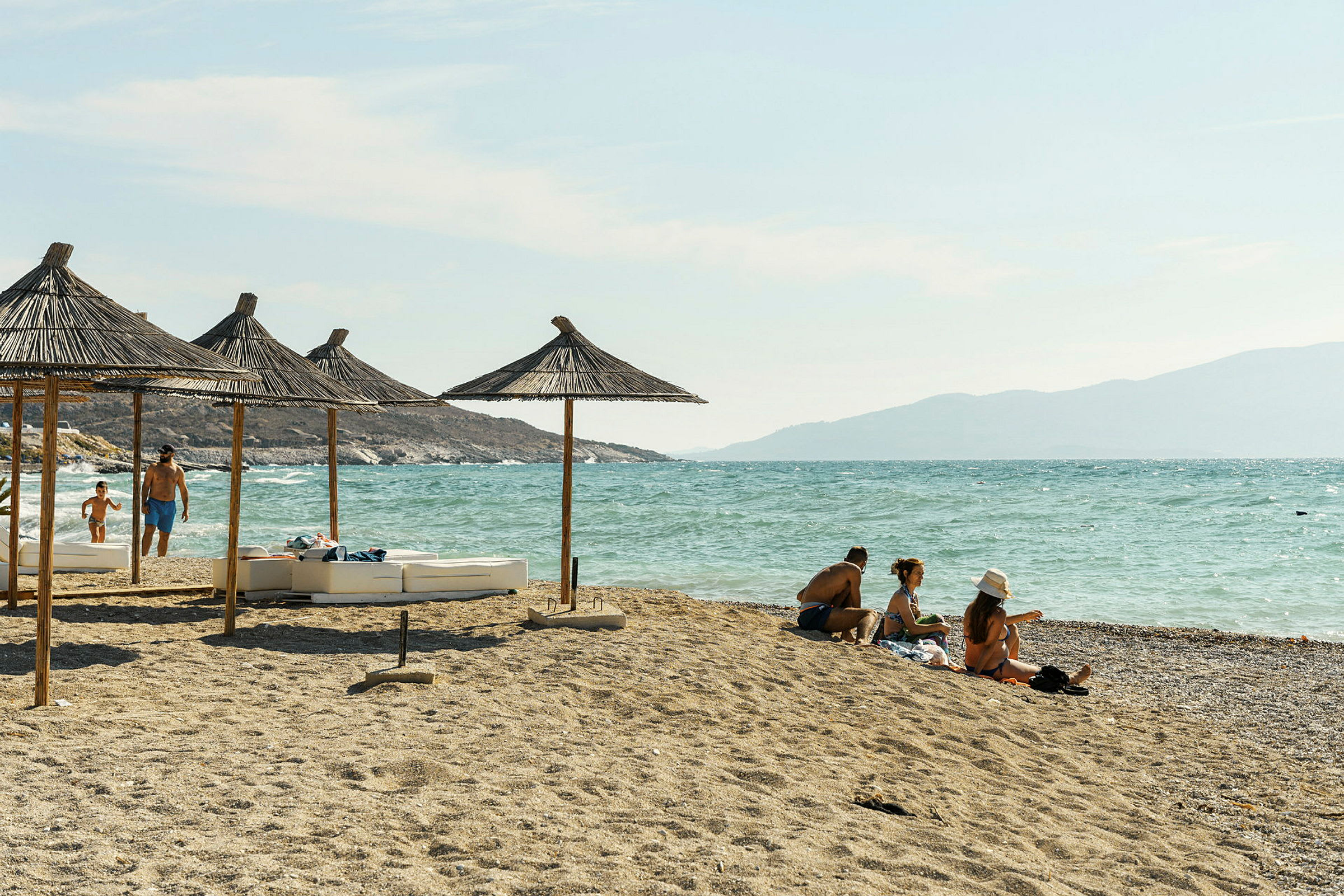 Strand med parasoller av halmtak og personer som nyter solen ved sjøen. (AI-generert tekst)