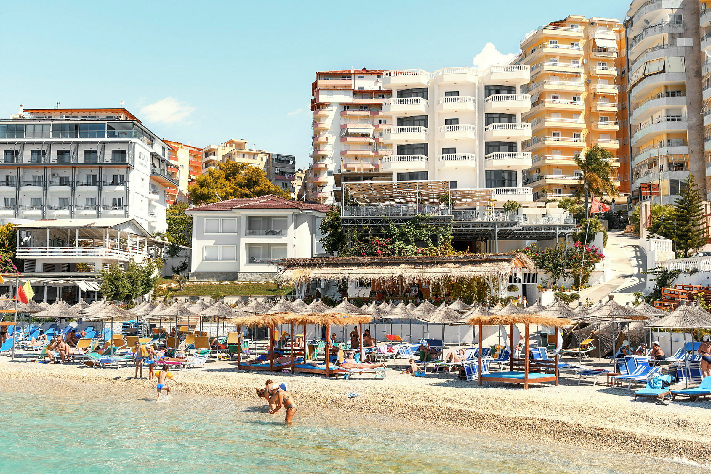 Strand med solsenger og parasoller foran bygninger. (AI-generert tekst)