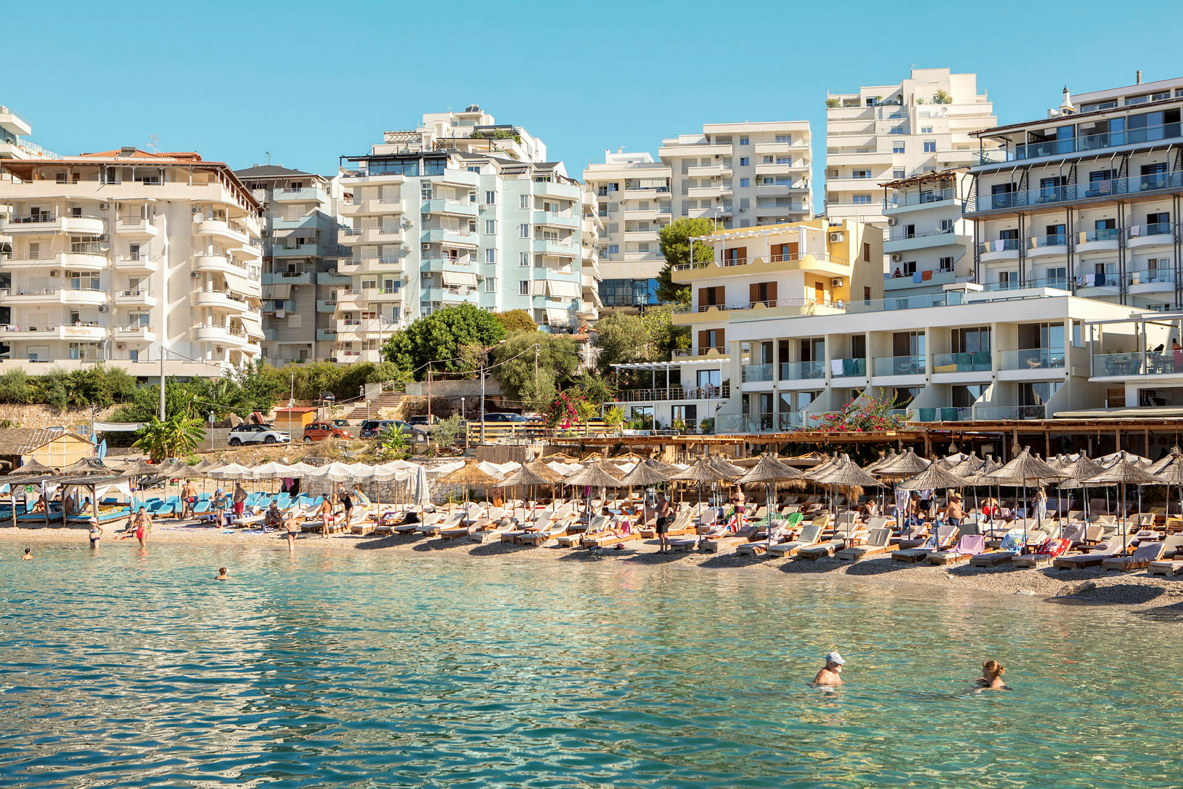En strand med solsenger og parasoller foran flere moderne bygninger. (AI-generert tekst)