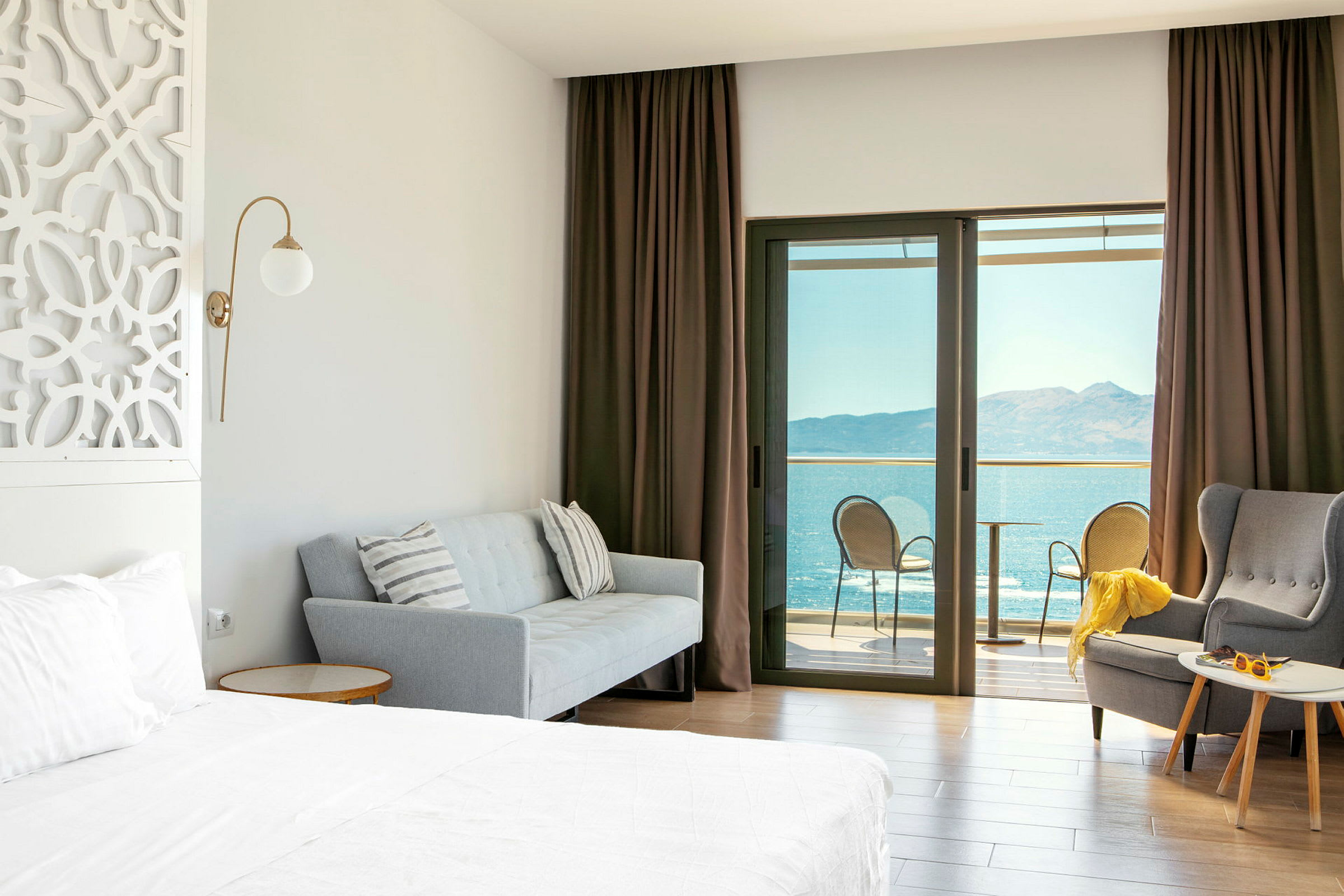 Hotellrom med seng, sofa, lenestol og balkong med havutsikt. (AI-generert tekst)