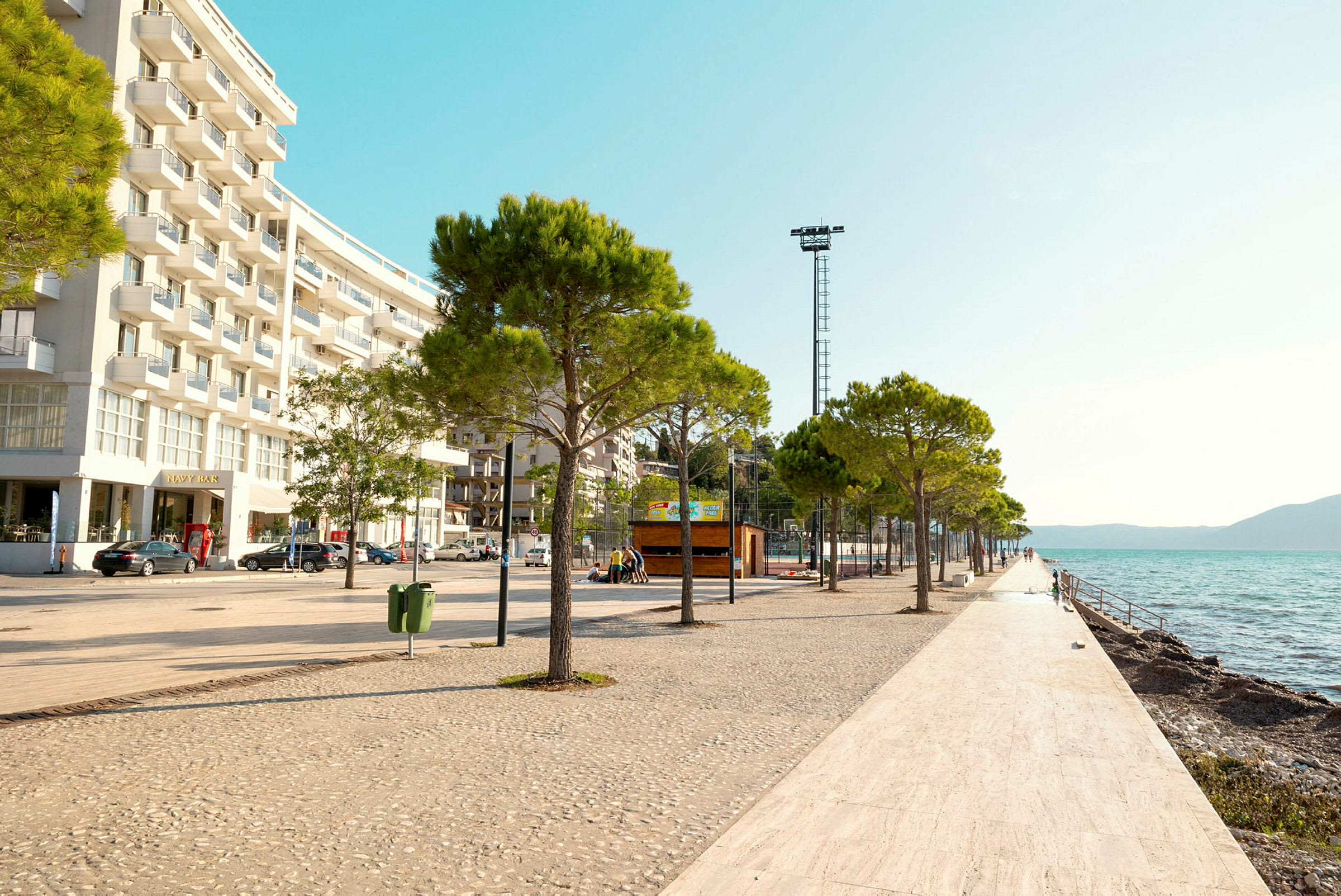 En strandpromenade med trær og hvitflislagt gulv langs vannet, med en moderne bygning til venstre. (AI-generert tekst)