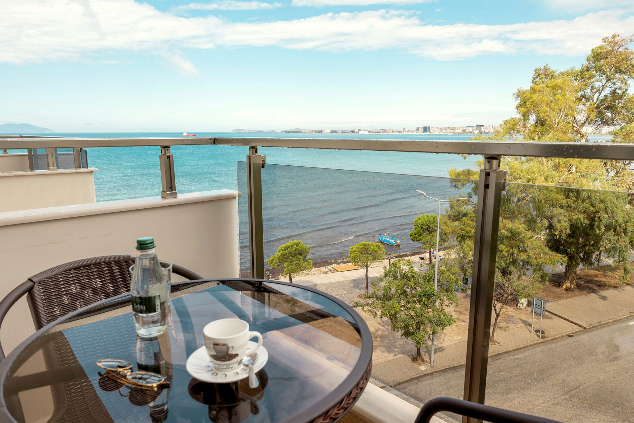 Utsikt fra en balkong med et lite bord hvor det står en kaffekopp, en flaske vann og et par solbriller. I bakgrunnen sees et hav og en strand med trær. (AI-generert tekst)