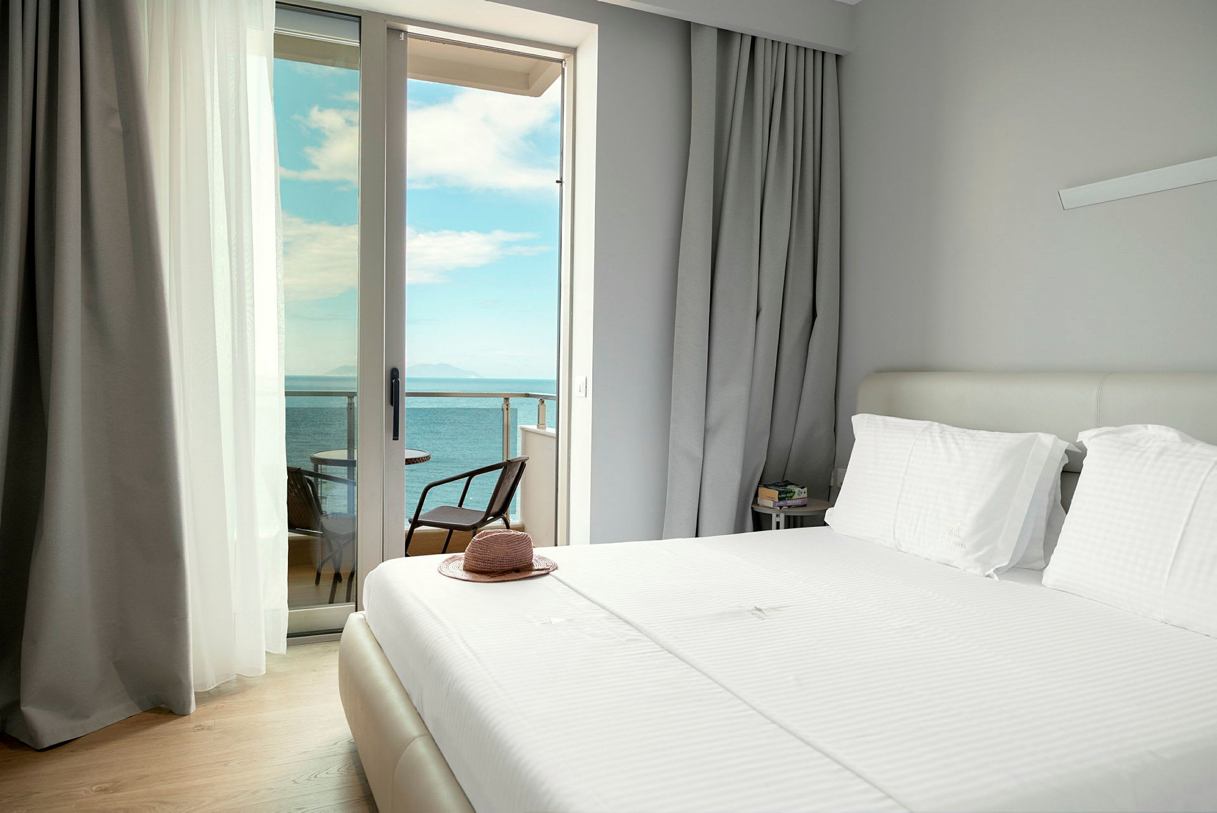 Et hotellrom med en oppredd seng og åpne gardiner som viser utsikt over havet fra balkongen. (AI-generert tekst)