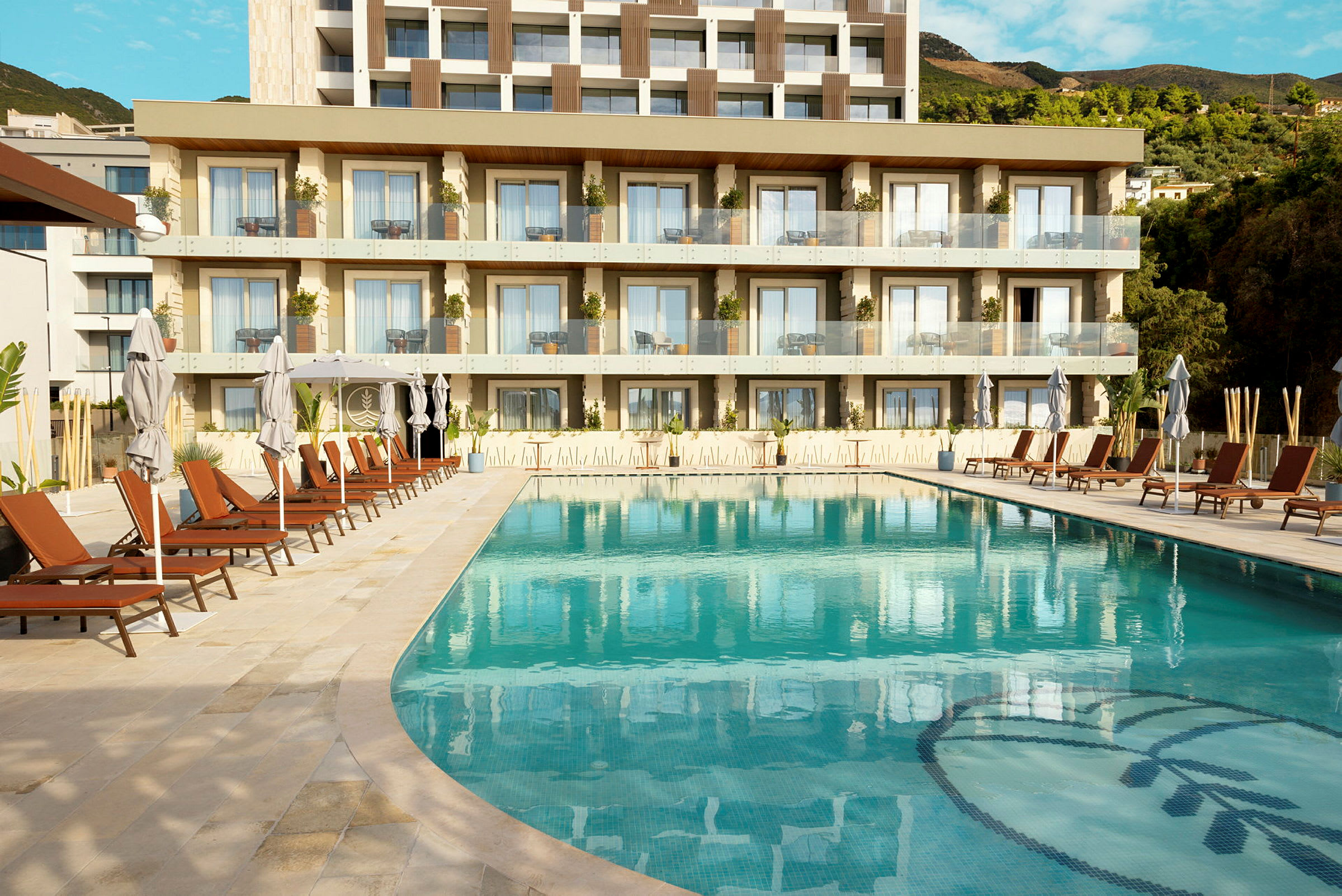 Bilde av et svømmebasseng med solsenger og parasoller foran et moderne hotell med balkonger. (AI-generert tekst)