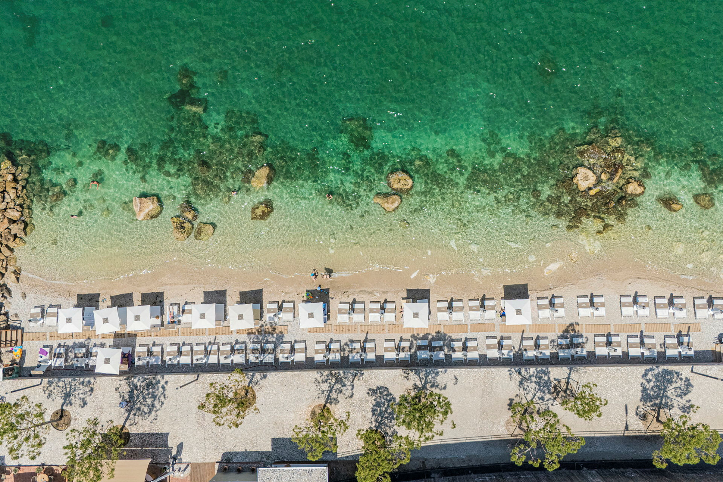Luftfoto av en strand med stoler og parasoller, grønt vann og små steiner synlige under overflaten. (AI-generert tekst)