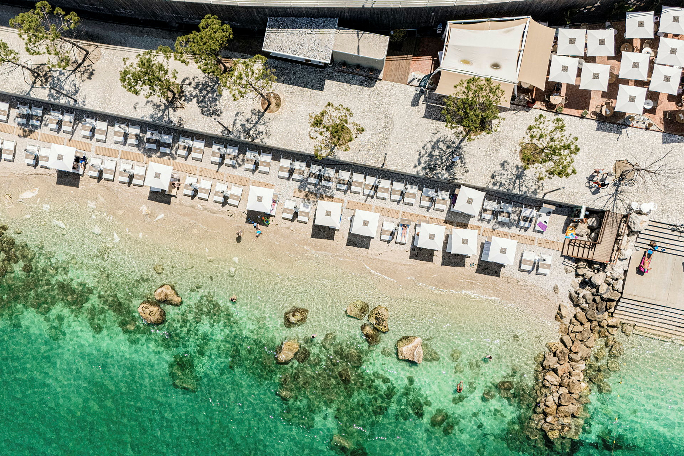 Flyfoto av en strand med parasoller og solsenger, samt klart, turkis vann. (AI-generert tekst)
