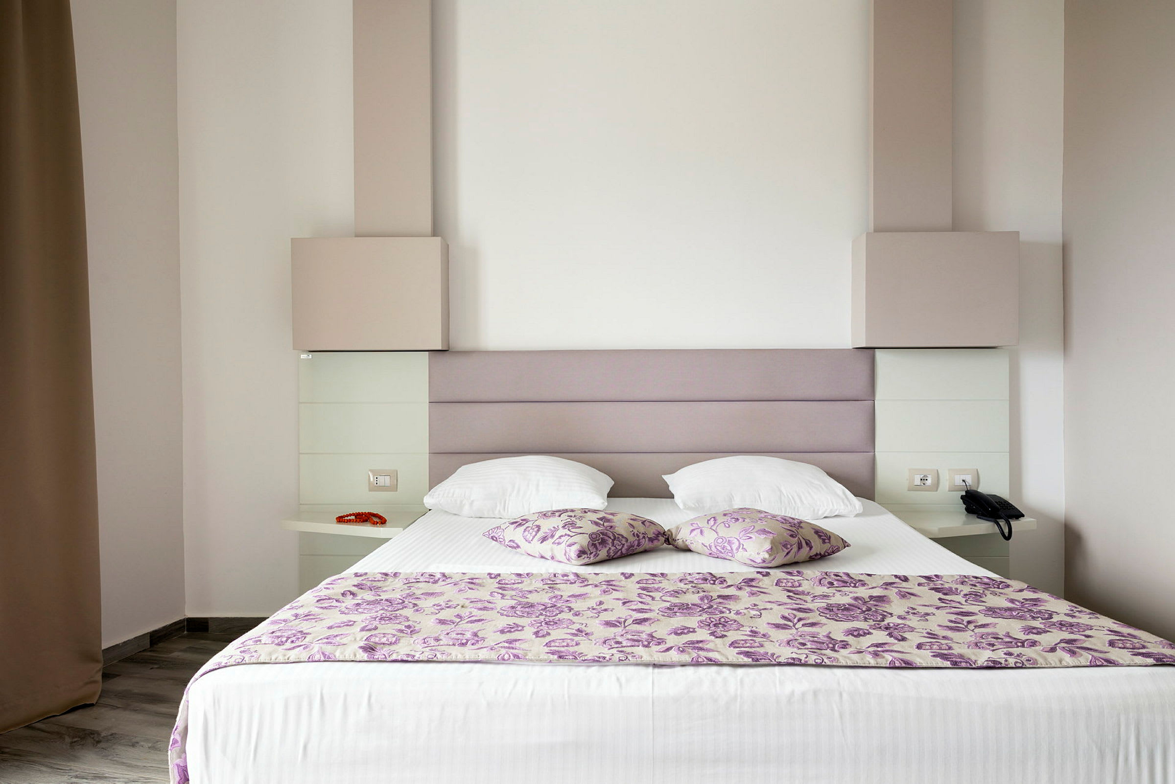 Et hotellrom med en oppredd seng dekorert med lilla blomstermønstrede puter og sengeteppe. (AI-generert tekst)