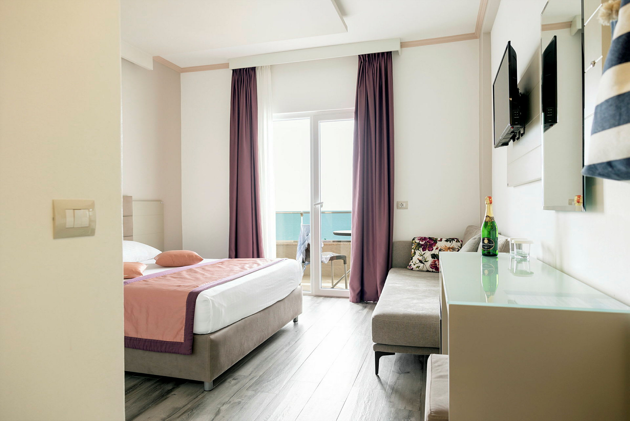 Hotellrom med en dobbeltseng, sofa, TV på veggen og glassdør som leder til en balkong. (AI-generert tekst)