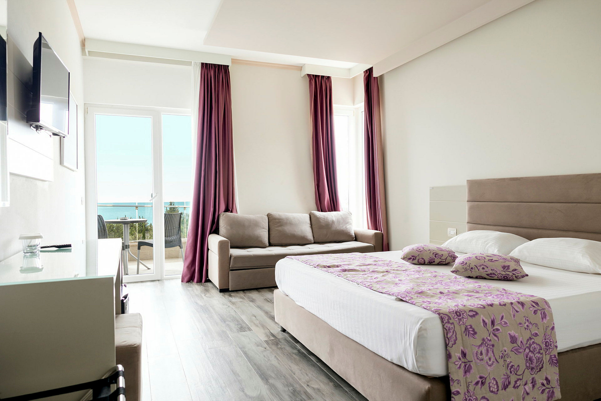 Et hotellrom med en dobbeltseng, en sofa og en balkong med utsikt mot havet. (AI-generert tekst)