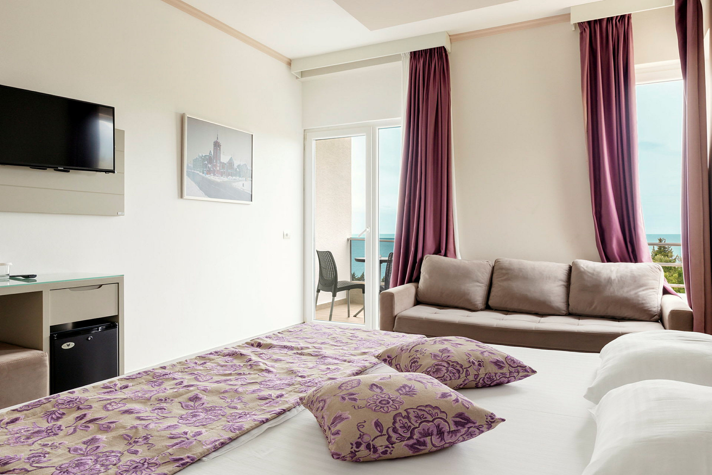 Hotellrom med seng, sofa, TV og en balkong med utsikt. (AI-generert tekst)