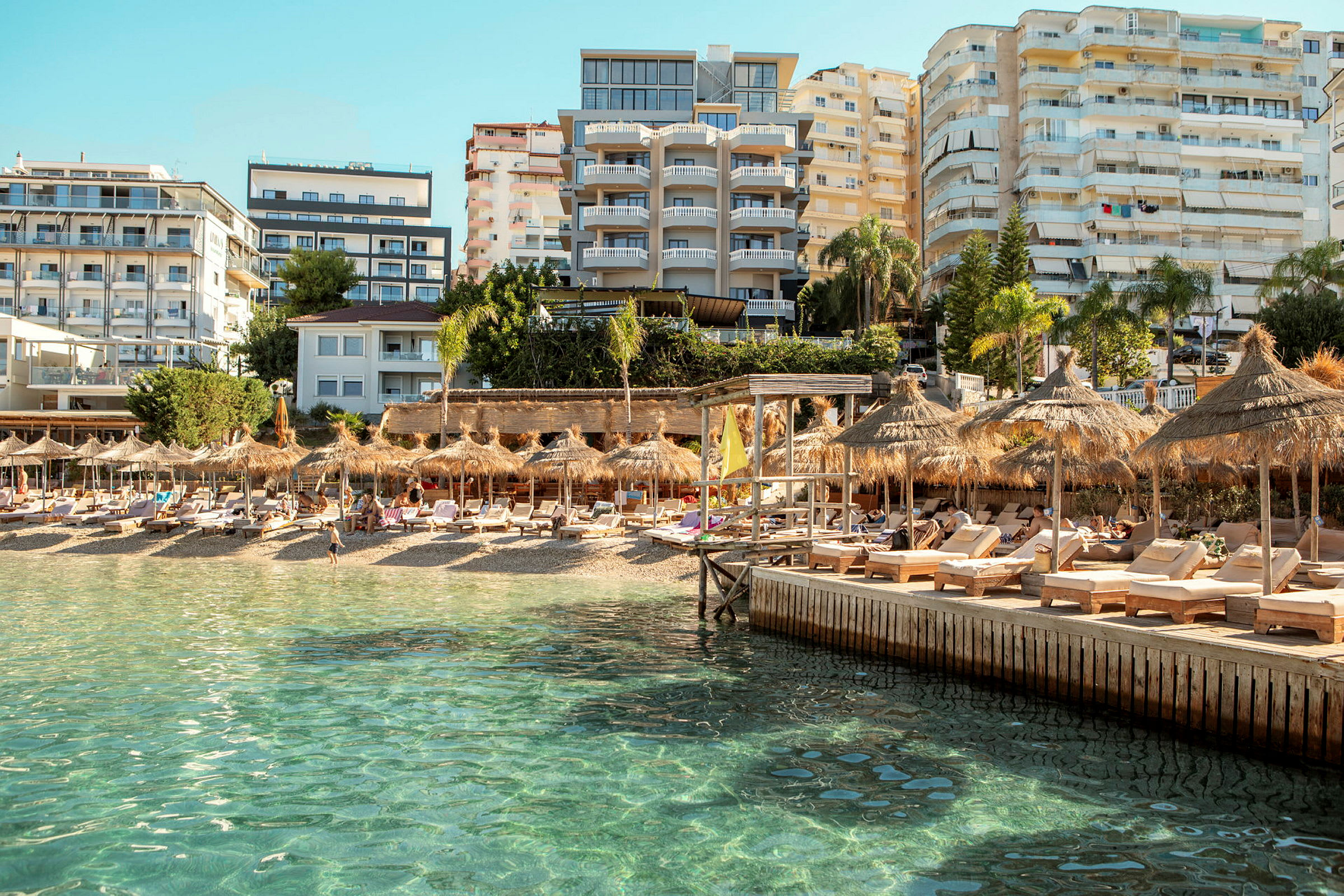 Strand med solsenger og parasoller, omgitt av hotellbygninger og en klarblå himmel. (AI-generert tekst)