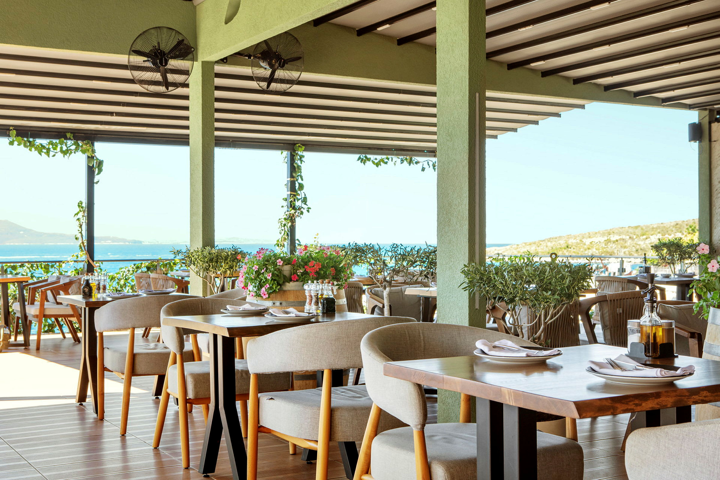 En restaurant med dekkede bord og utsikt over havet, dekorert med grønne planter. (AI-generert tekst)
