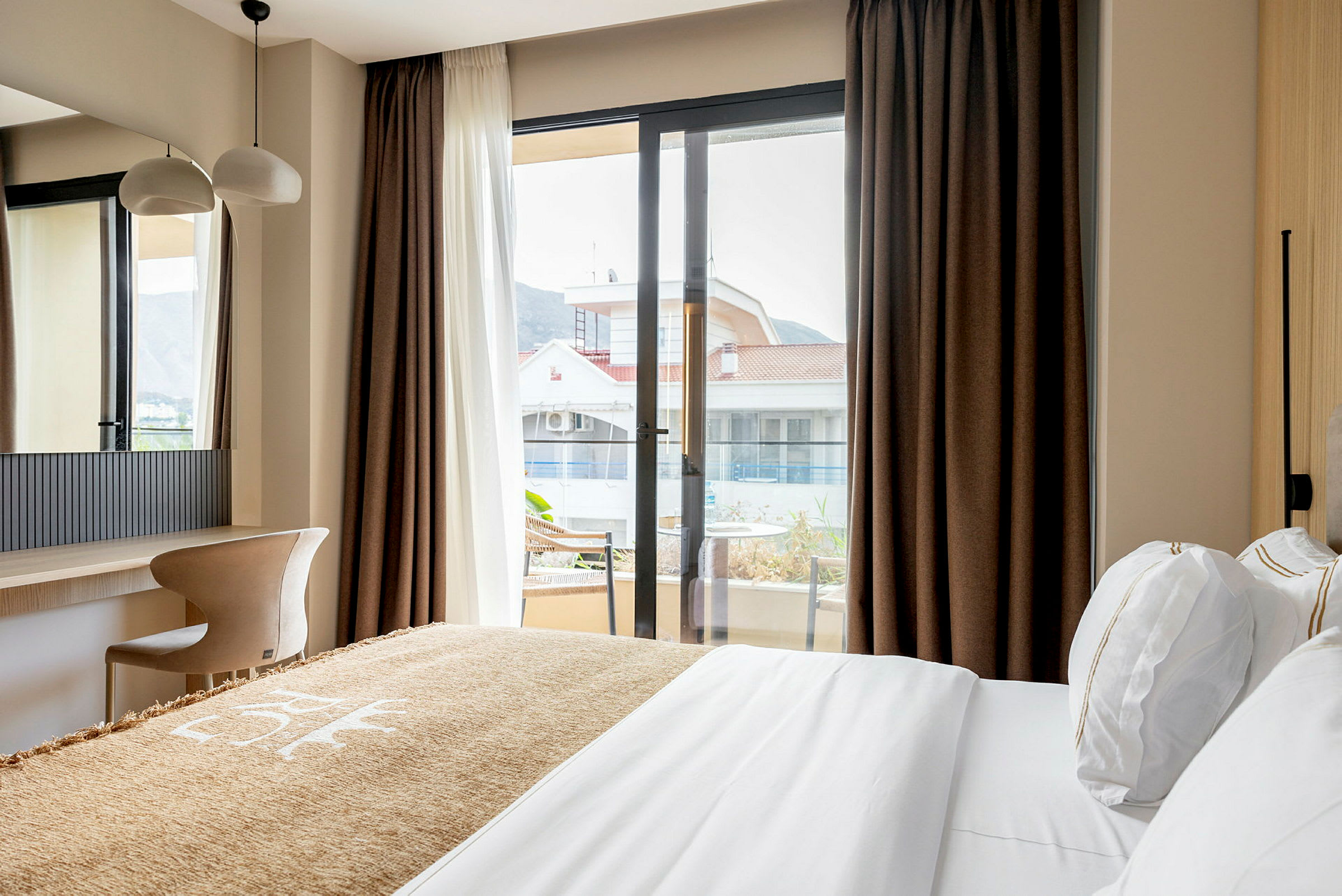 Et hotellrom med en seng, skrivebord og stol, samt utsikt mot en bygning gjennom et stort vindu. (AI-generert tekst)