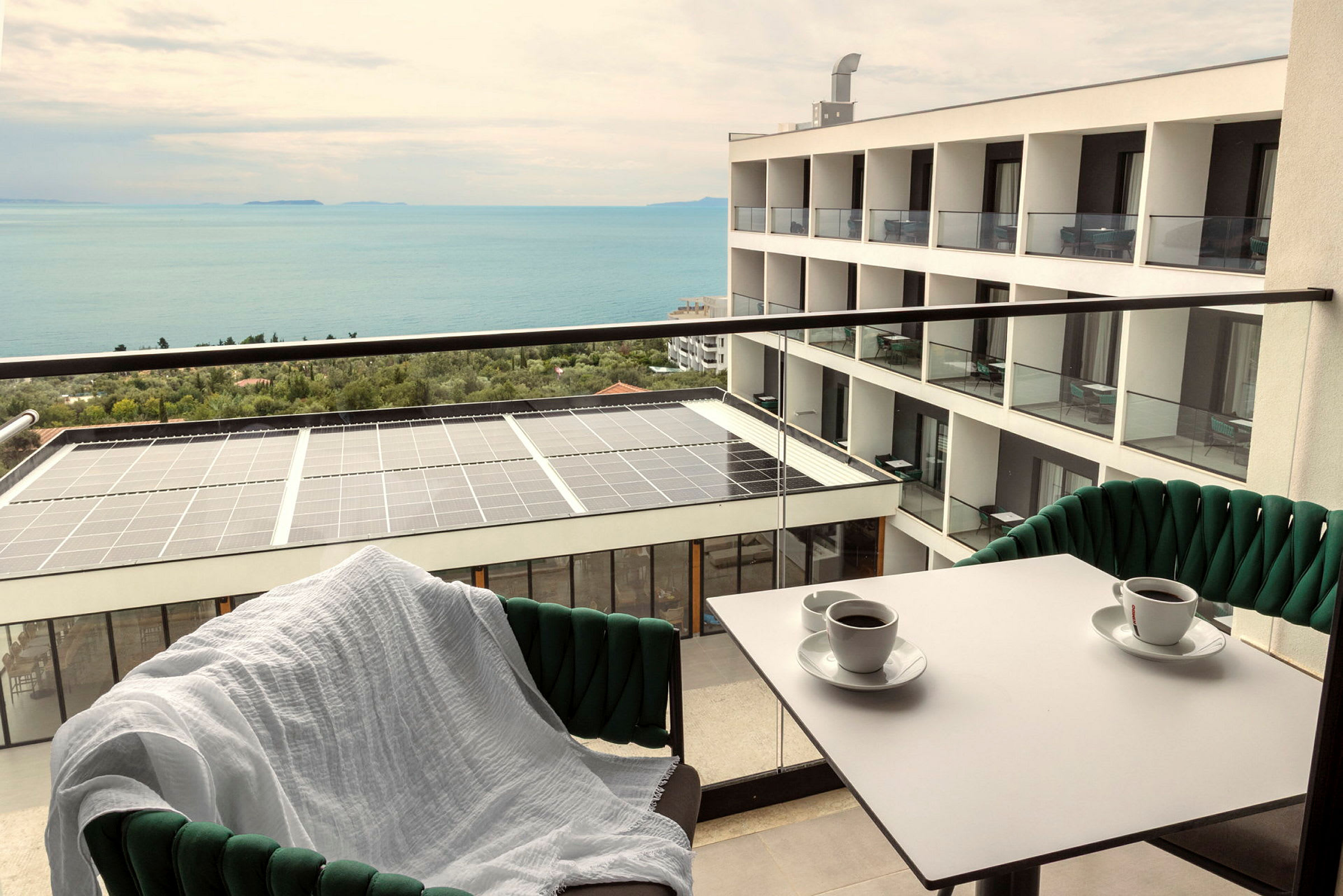 Utsikt fra en balkong med to kaffekopper på bordet, en stol med hvitt teppe, en bygning og havet i bakgrunnen. (AI-generert tekst)