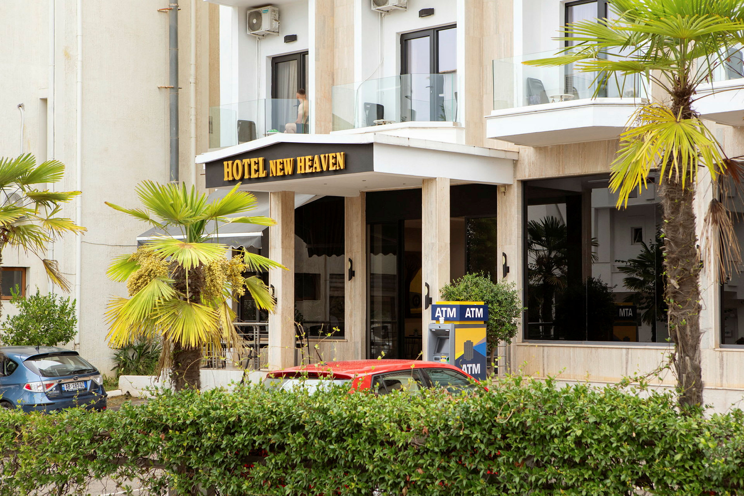 Bygningens front med teksten 'Hotel New Heaven', omgitt av palmer og en minibank. (AI-generert tekst)