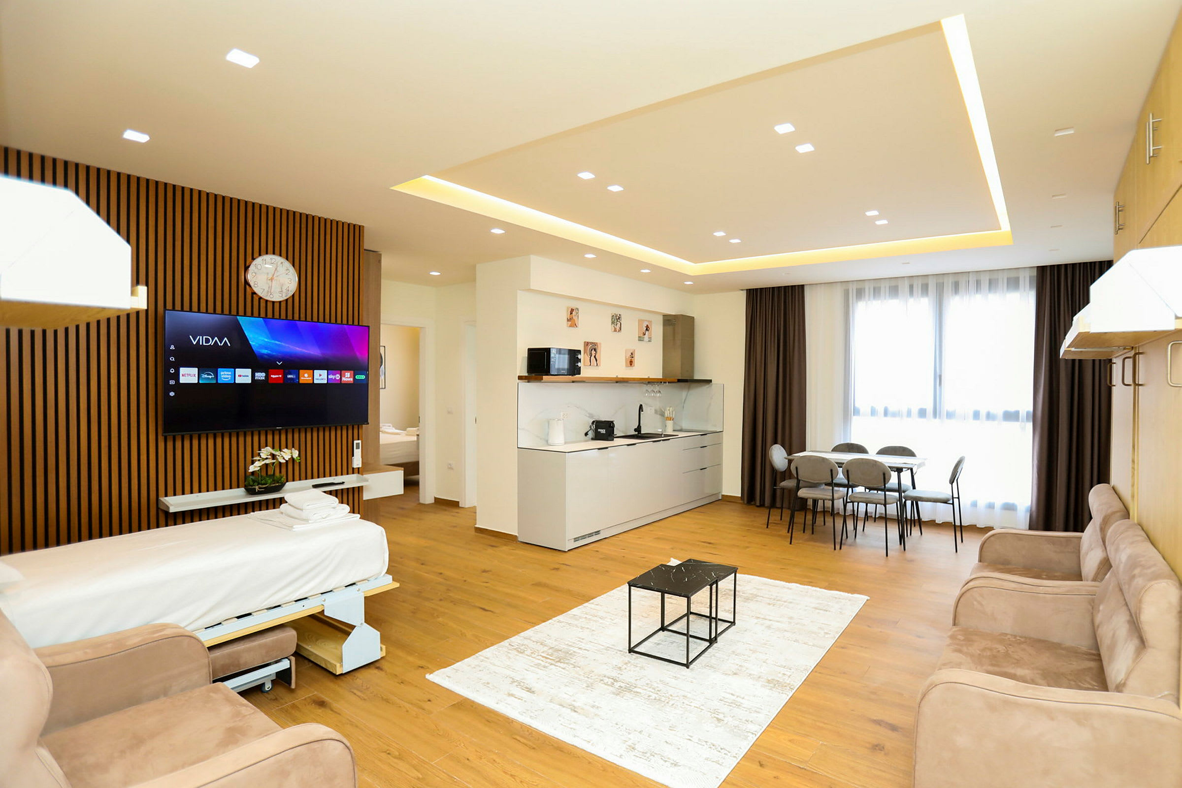 Et moderne hotellrom med åpen planløsning som inkluderer en sovesofa, TV, kjøkkenområde, spisebord og store vinduer med gardiner. (AI-generert tekst)