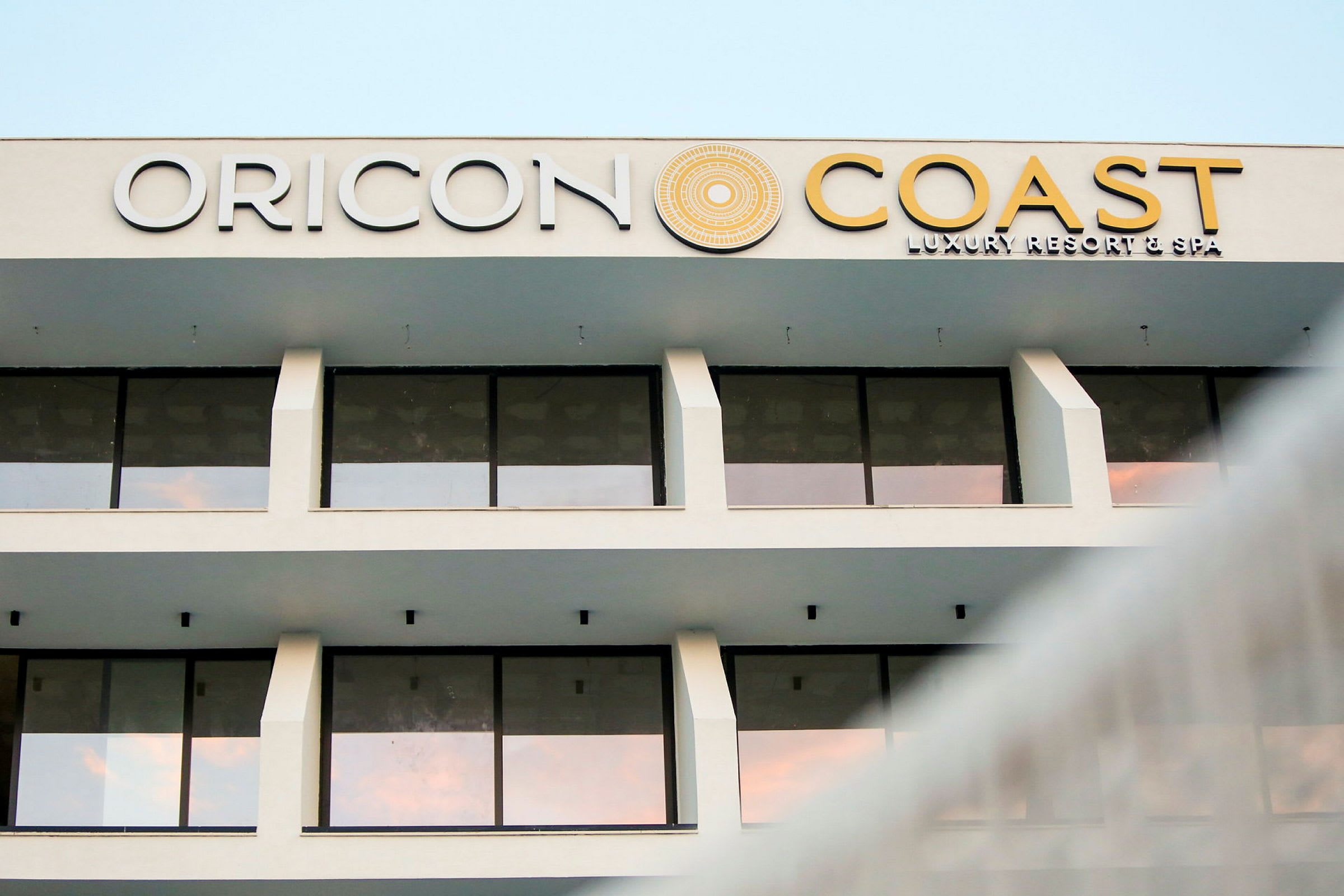 Fasaden til en hotellbygning med store glassvinduer og teksten "Oricon Coast Luxury Resort & Spa" på toppen. (AI-generert tekst)